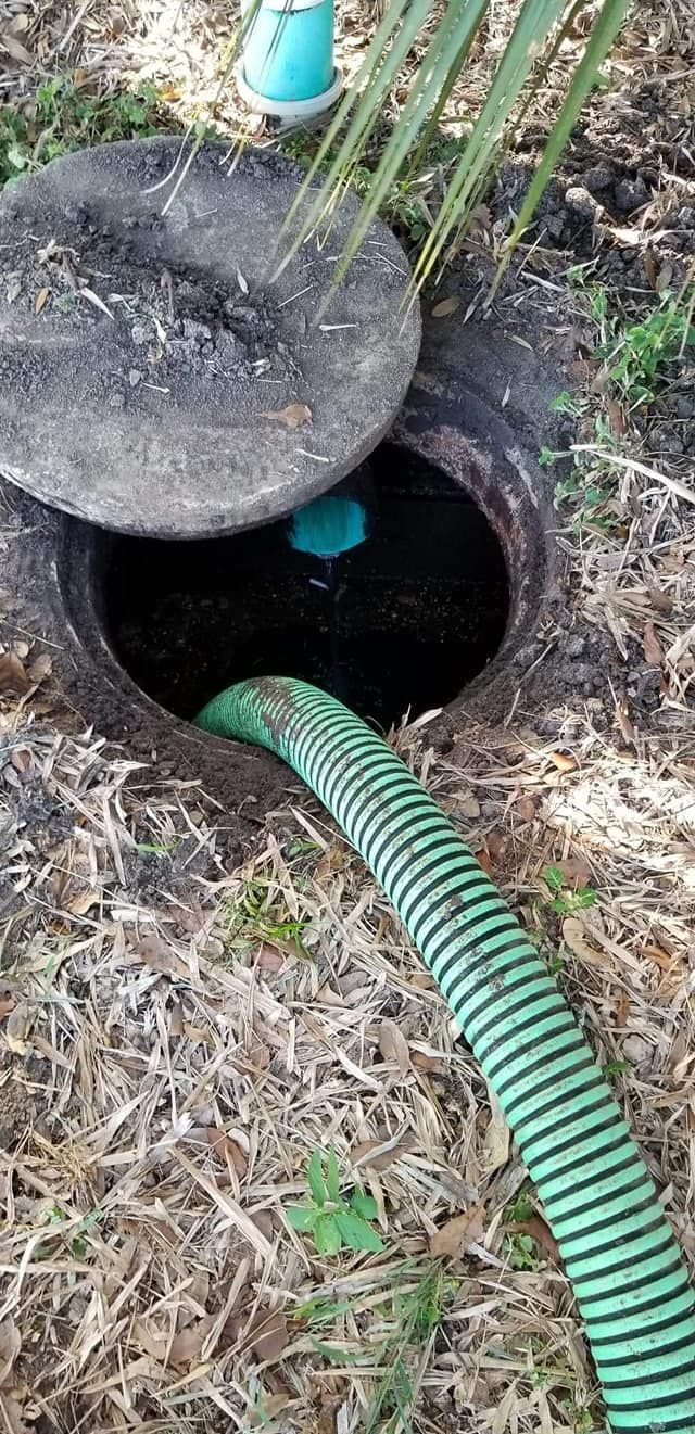 Open septic tank with a green hose, lid partially removed, surrounded by dry grass.