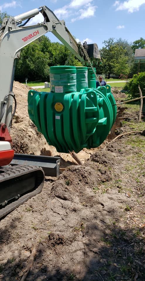 A green septic tank being lowered into a trench by a small excavator on a sunny day.