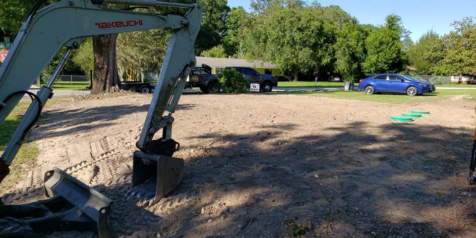 An excavator is in a gravel yard with a blue car and a house in the background.