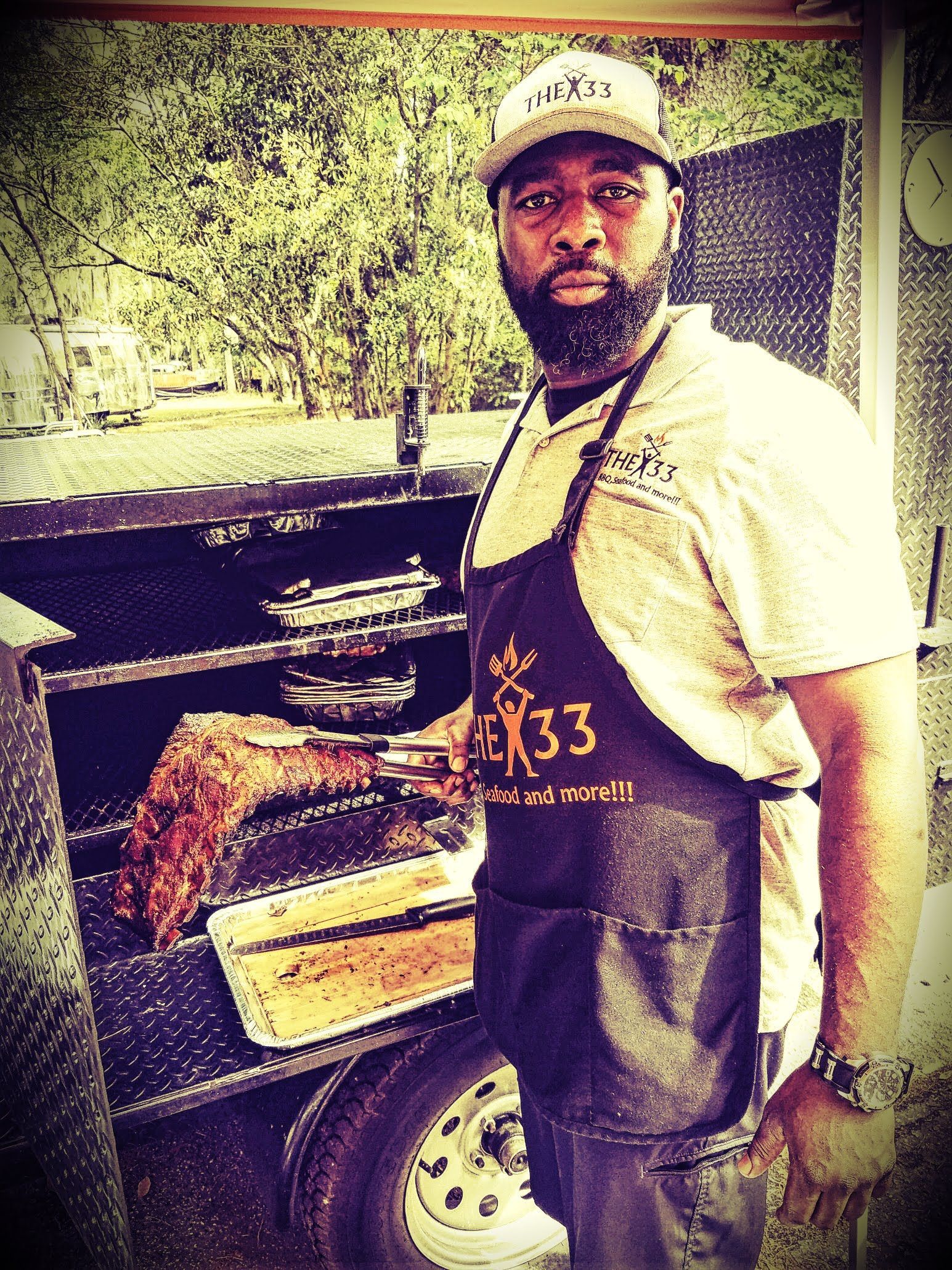 A person wearing a cap and an apron stands outdoors, using tongs to turn meat on a large barbecue smoker.