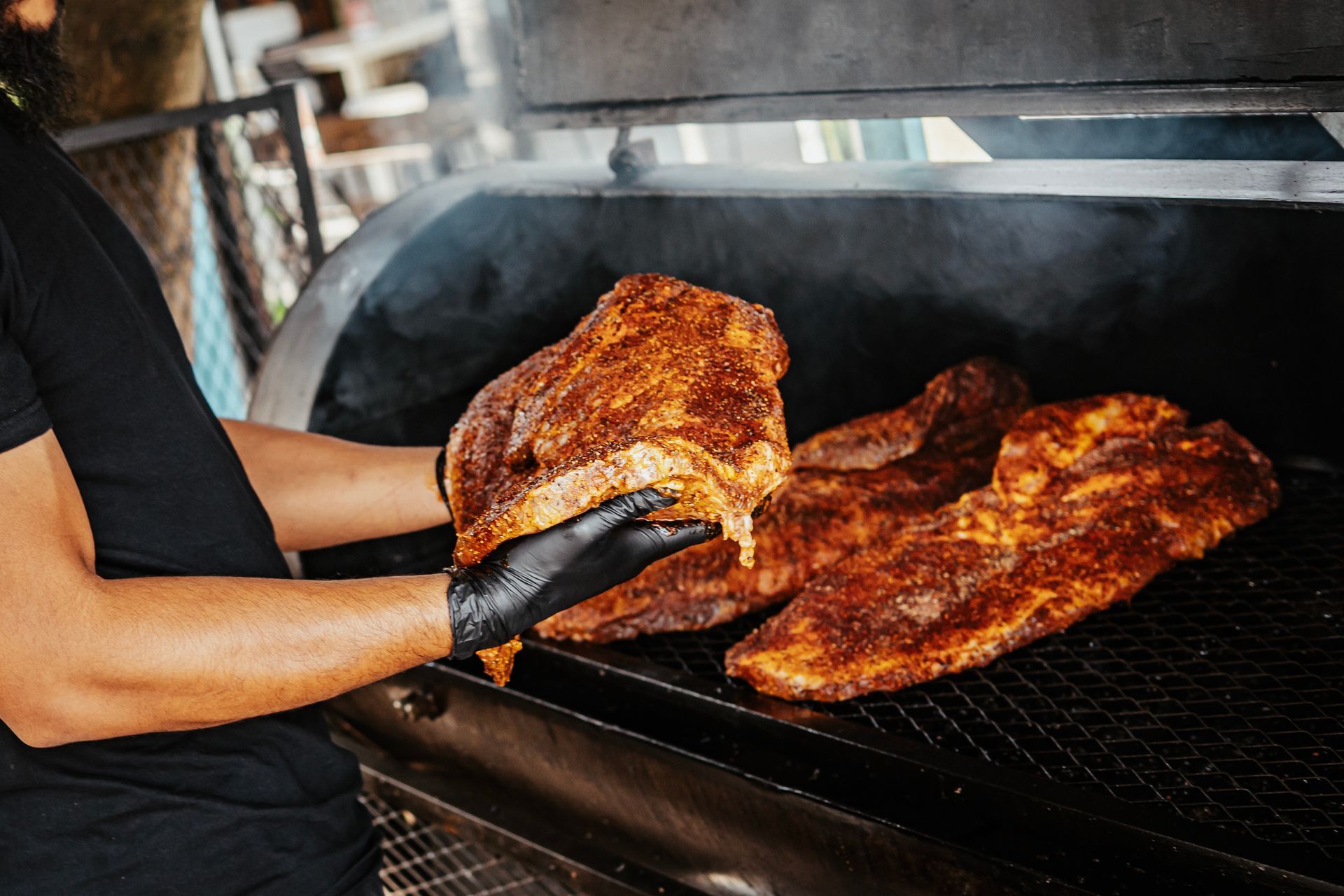A person wearing a black glove places a seasoned slab of meat onto the grate inside a large, smoking barbecue smoker.