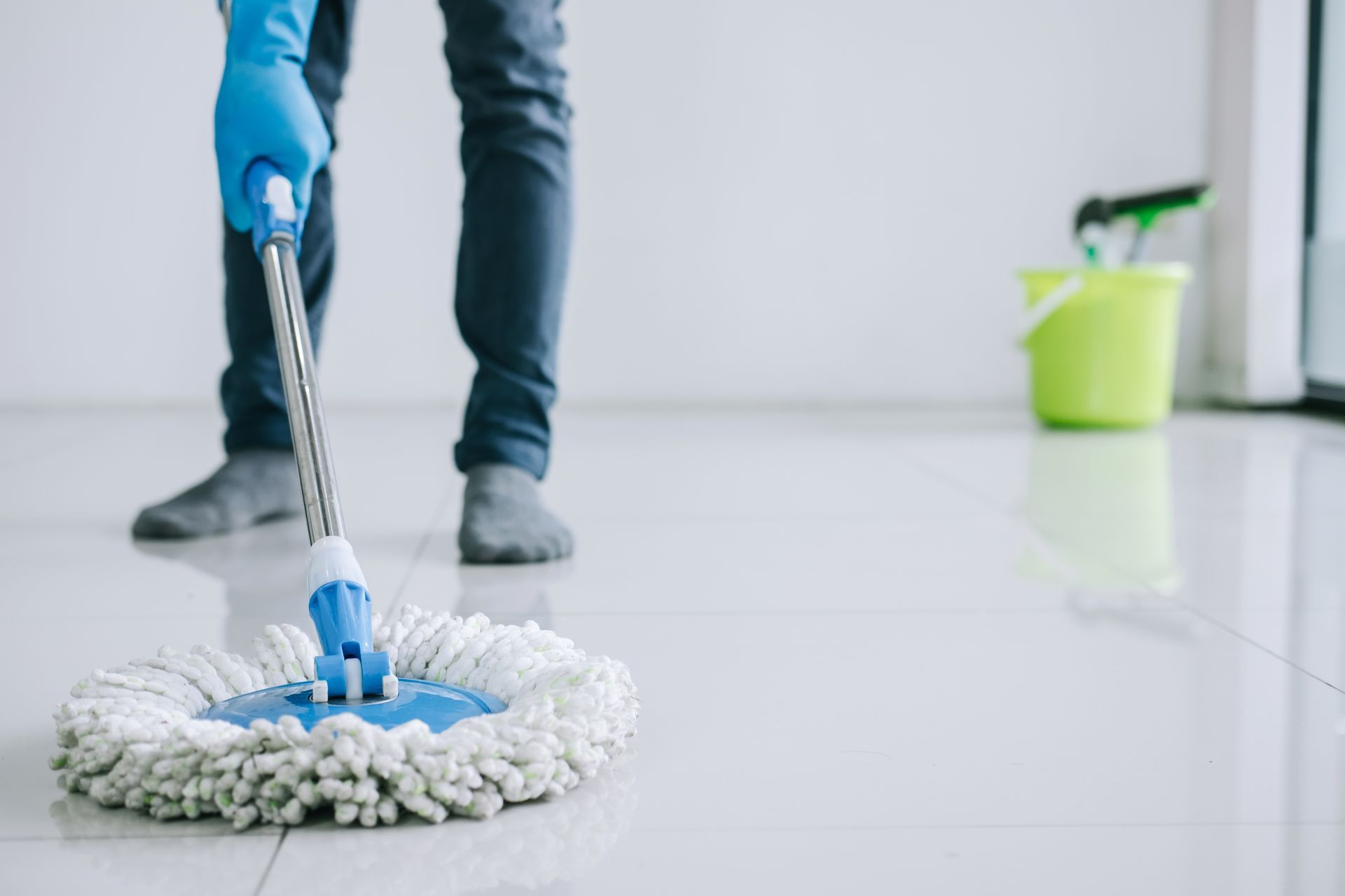 Person mopping a light-colored floor with a blue mop, wearing gloves and dark pants, with a bucket in the background.