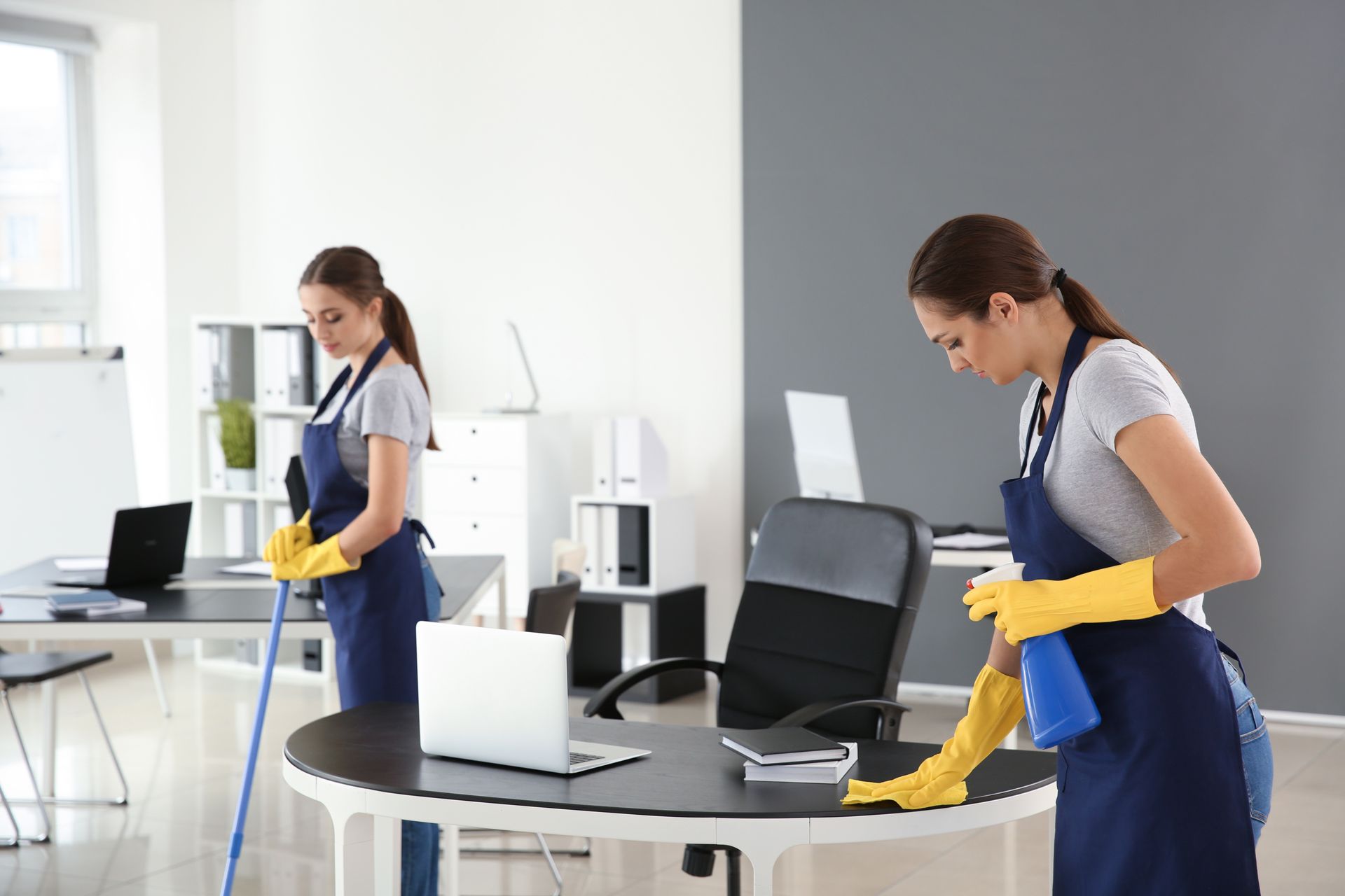 Two people cleaning an office: one mopping, the other wiping a desk, both wearing aprons and gloves.