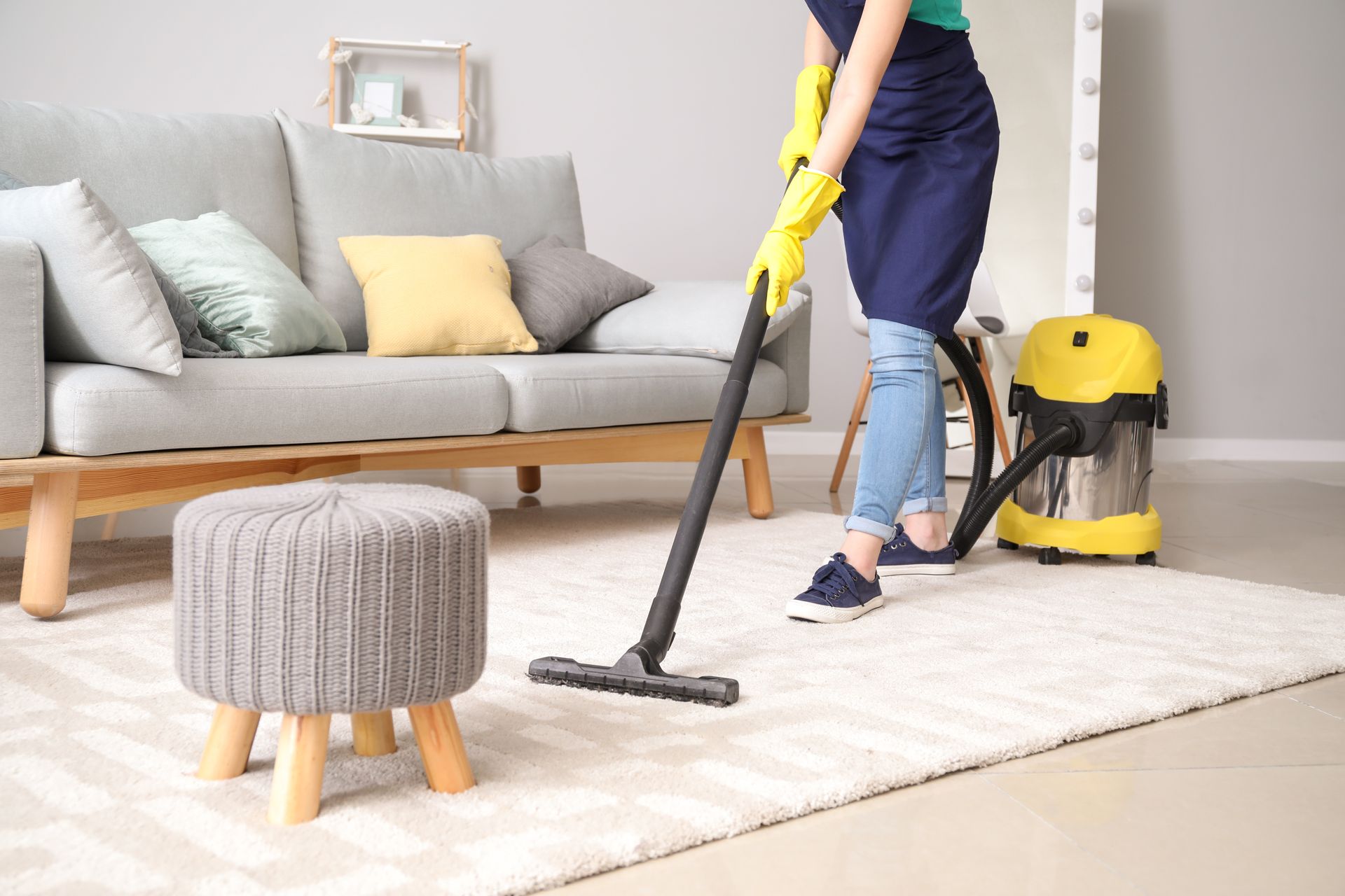 Person vacuuming a light-colored rug in a living room with a sofa, stool, and yellow vacuum cleaner.