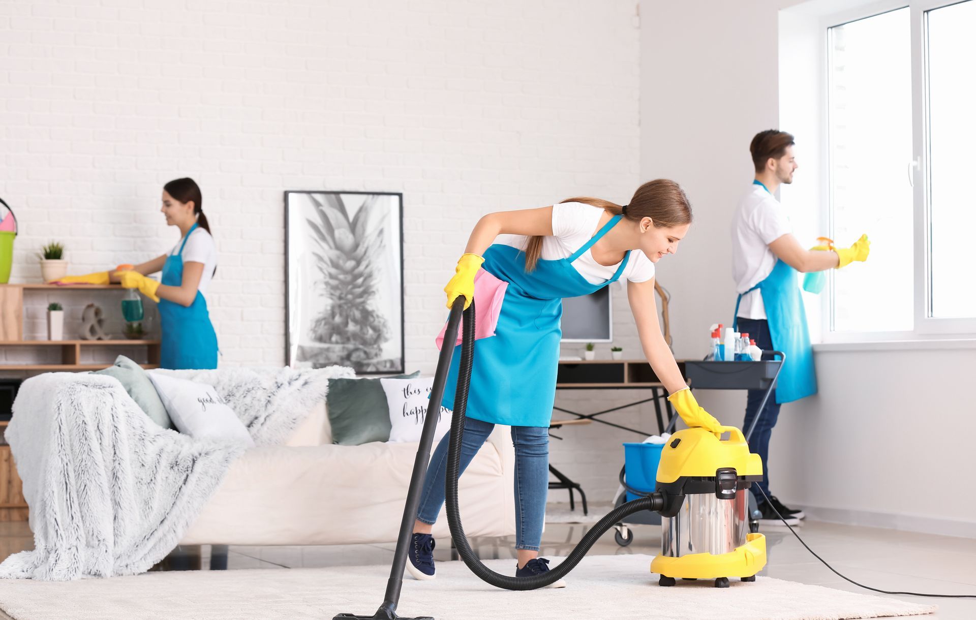 Three cleaners vacuum and dust in a bright living room with a sofa and large window.
