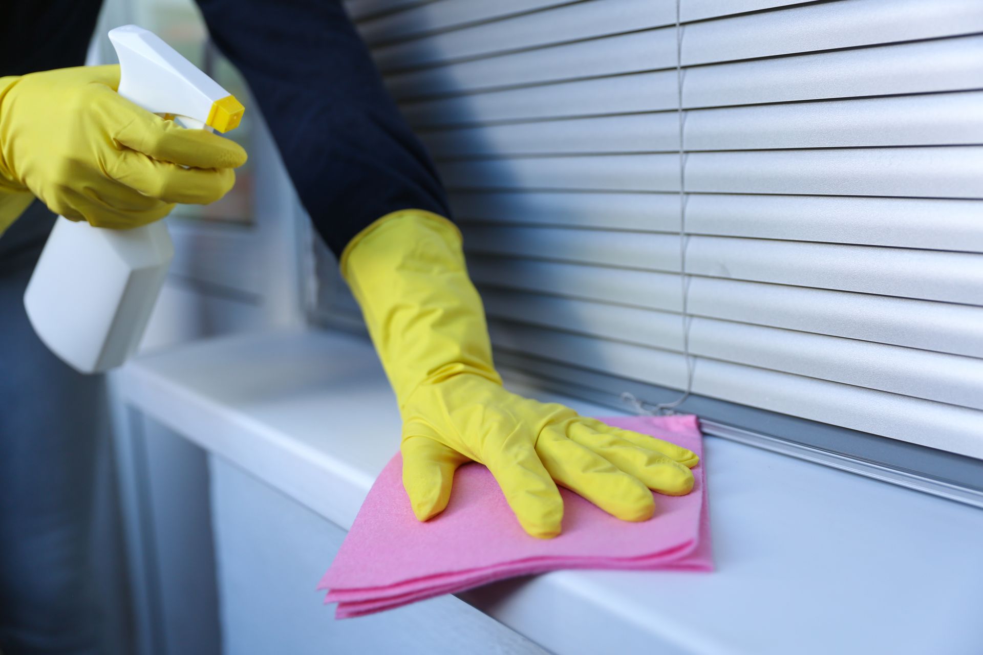 Gloved hands cleaning a window sill with a pink cloth and spray bottle