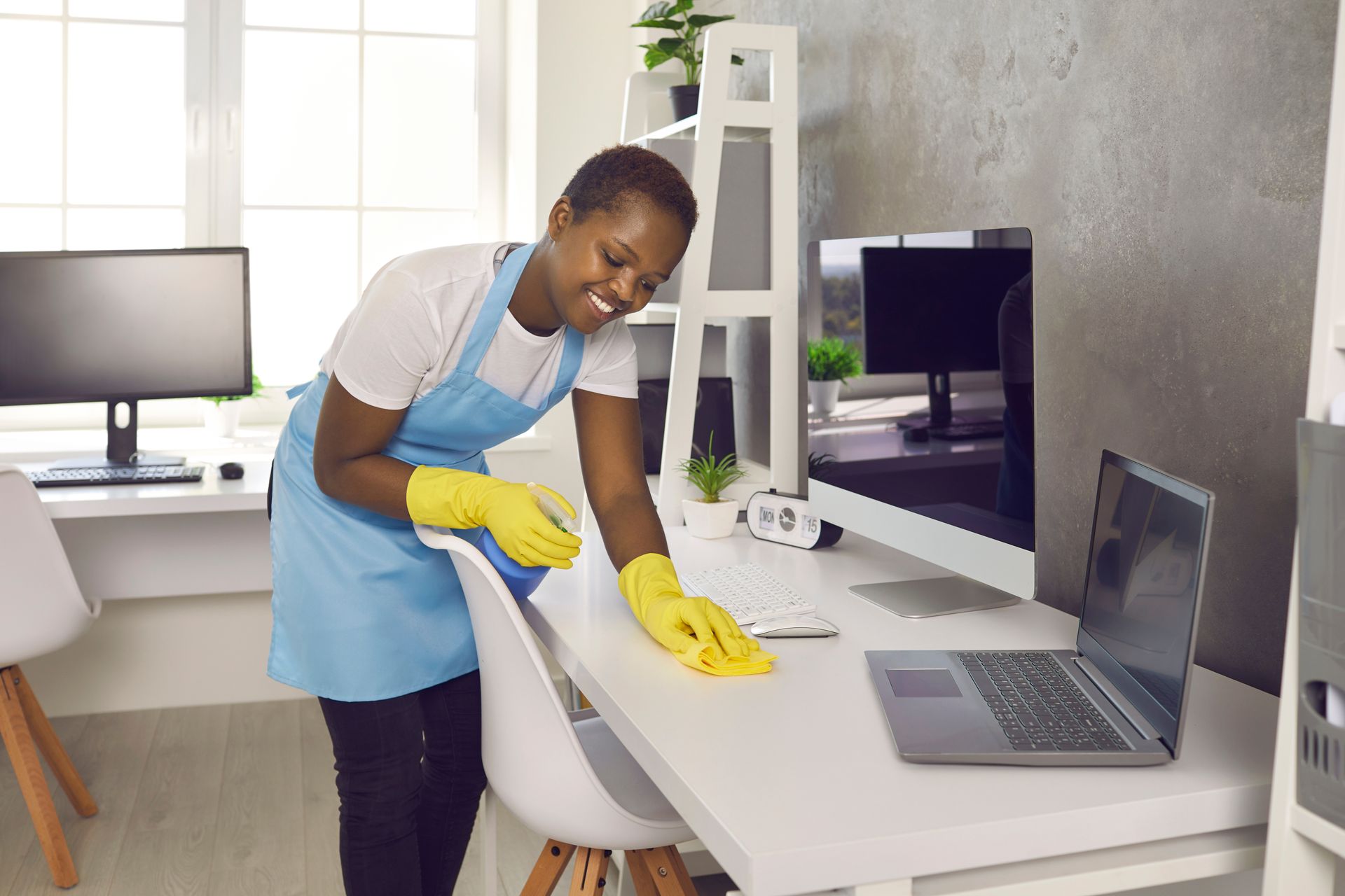 Woman in blue apron and yellow gloves cleaning a white desk in an office. Woman in blue apron and yellow gloves cleaning a white desk in an office.