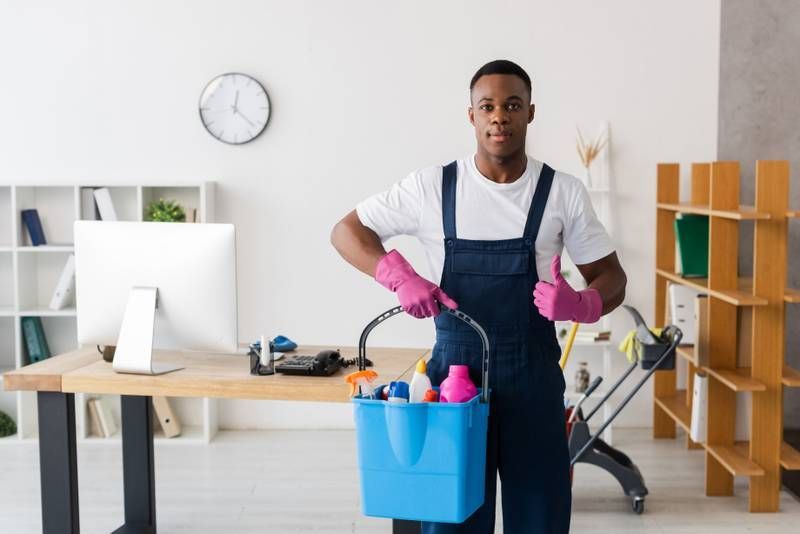 Man in overalls, pink gloves, holding cleaning supplies bucket, thumbs up, office setting.