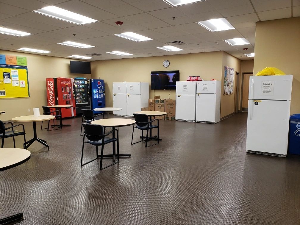 A break room with vending machines, tables, chairs, refrigerators, and a clock.
