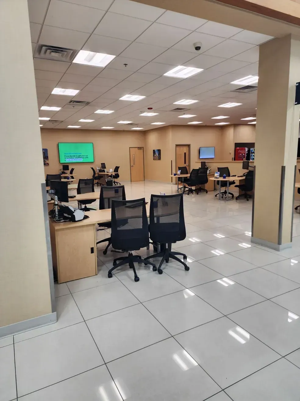 Interior of an office with desks, chairs, and monitors. Beige walls, white tiled floor, and overhead lighting.