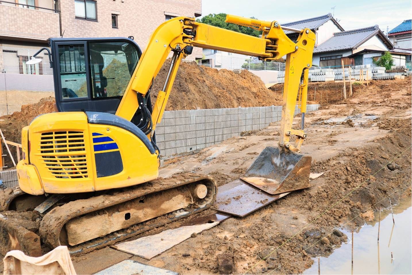 Yellow excavator working on a muddy construction site beside houses and a trench