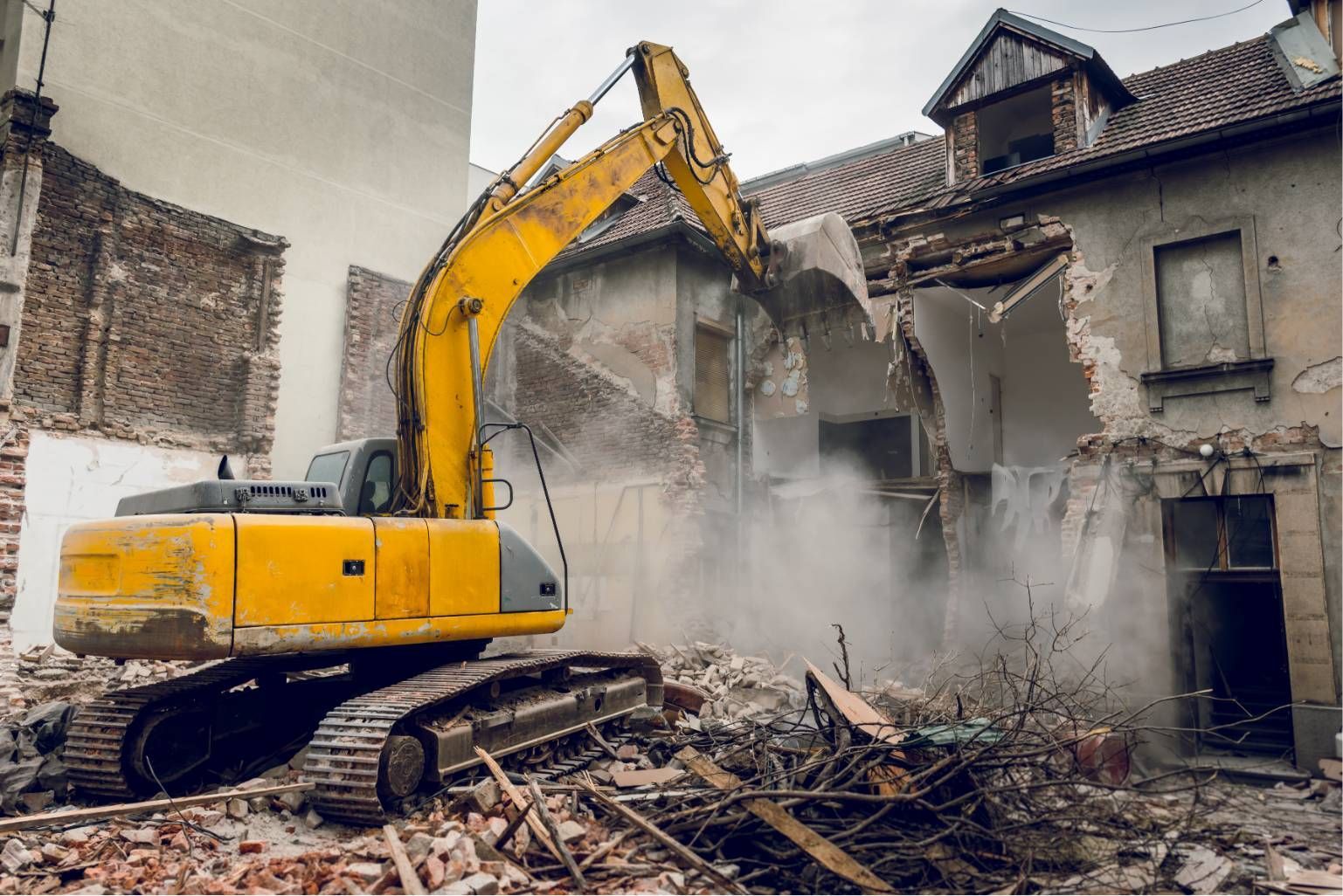 Yellow excavator demolishing a damaged house amid rubble and dust