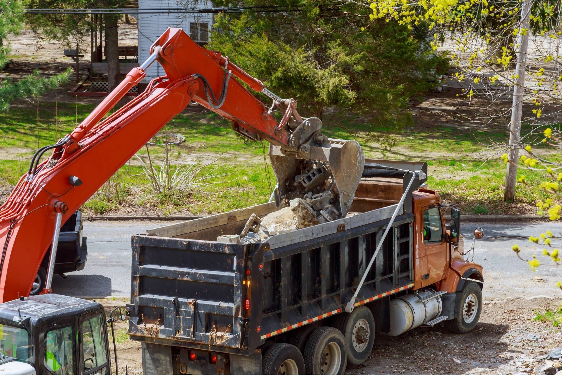 Orange excavator loading dirt into a black dump truck at a roadside construction site