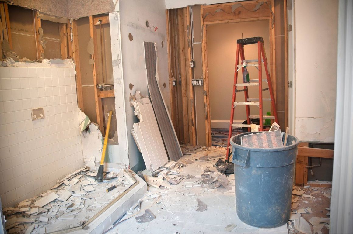 Demolition site with exposed studs, debris on floor, a blue trash bin, and a red ladder in a doorway