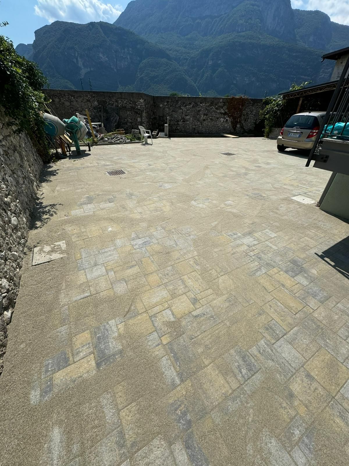 Un cortile lastricato in pietra con un'auto parcheggiata, circondato da un basso muro di pietra con le montagne sullo sfondo.
