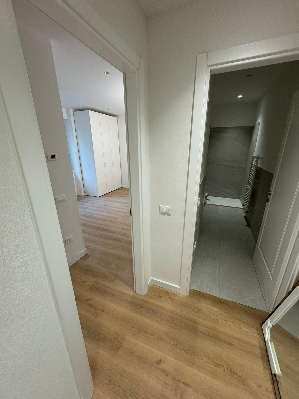Un corridoio con pavimento in legno chiaro conduce a una stanza con un armadio bianco, e una porta adiacente rivela un bagno.