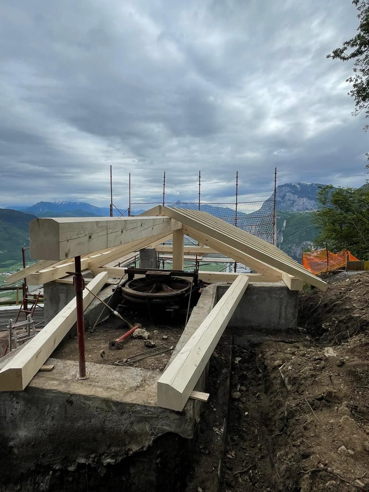 Una struttura di tetto in legno in costruzione su fondamenta in cemento, con vista su un paesaggio montuoso sotto un cielo nuvoloso.
