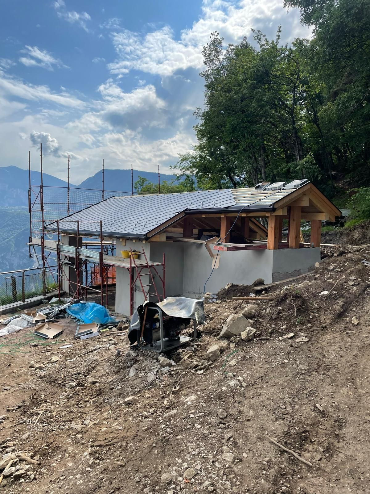 Un piccolo edificio in costruzione con il tetto di tegole grigie sorge su un pendio roccioso con vista sulle montagne.