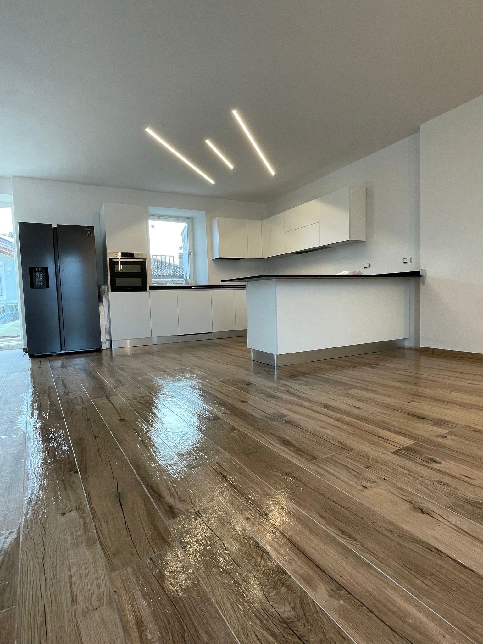 Cucina moderna con mobili bianchi, frigorifero nero e pavimento in legno lucido illuminato da faretti lineari a soffitto.