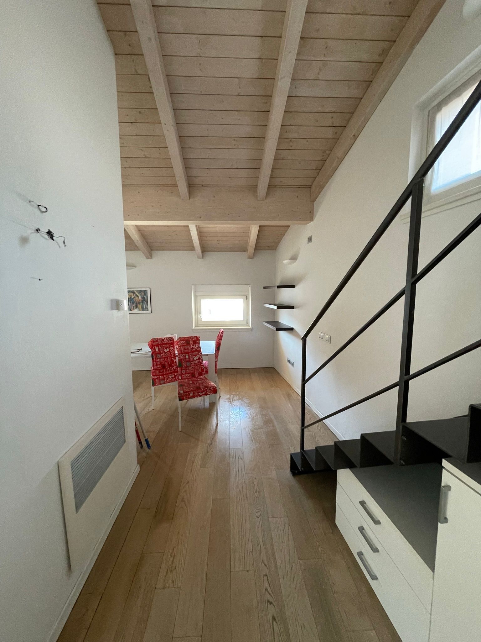 Una stanza luminosa con pavimento in legno e soffitto con travi a vista in legno chiaro, arredata con un tavolo da pranzo e una scala in metallo nero.