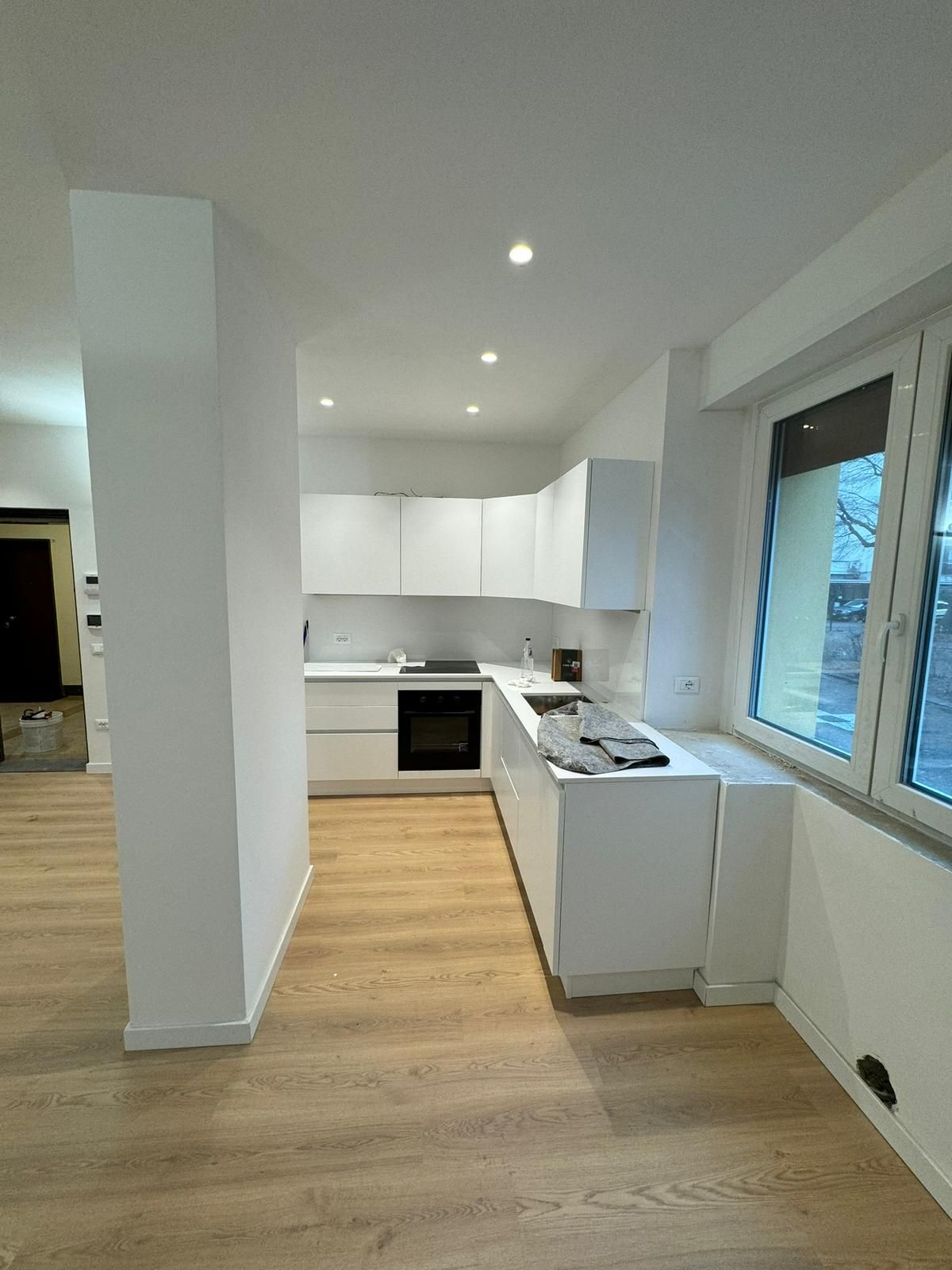 Una cucina moderna con mobili bianchi, pavimento in legno chiaro e un'isola affacciata su una finestra.