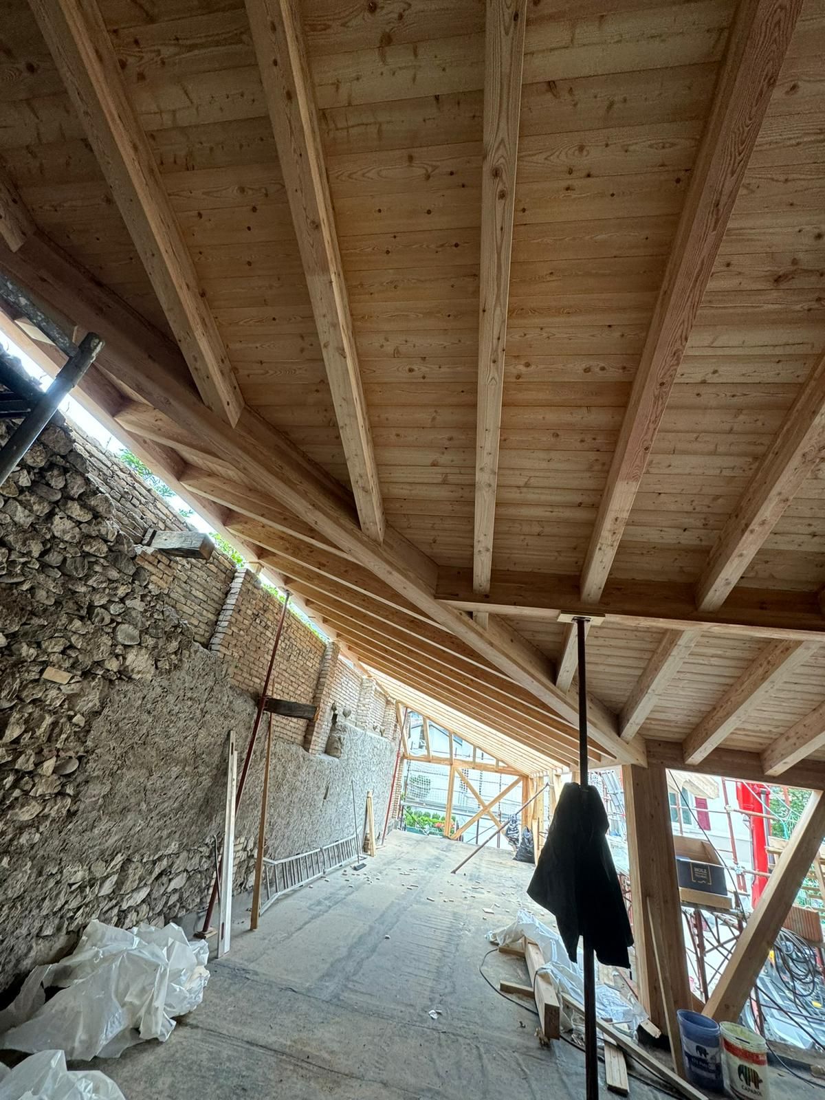 Vista interna che mostra la struttura di un tetto in legno in costruzione, con un muro in pietra sulla sinistra e un'impalcatura sottostante.