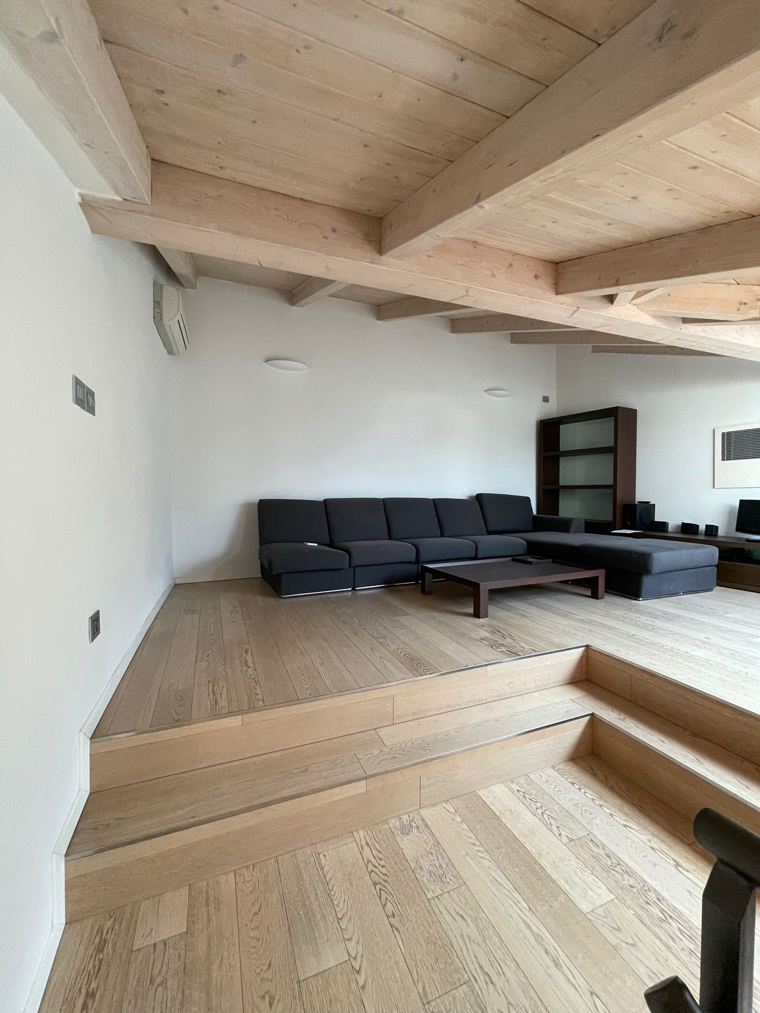 Una zona giorno con un divano scuro su una piattaforma di legno rialzata, caratterizzata da travi in ​​legno chiaro e una scala a più livelli.