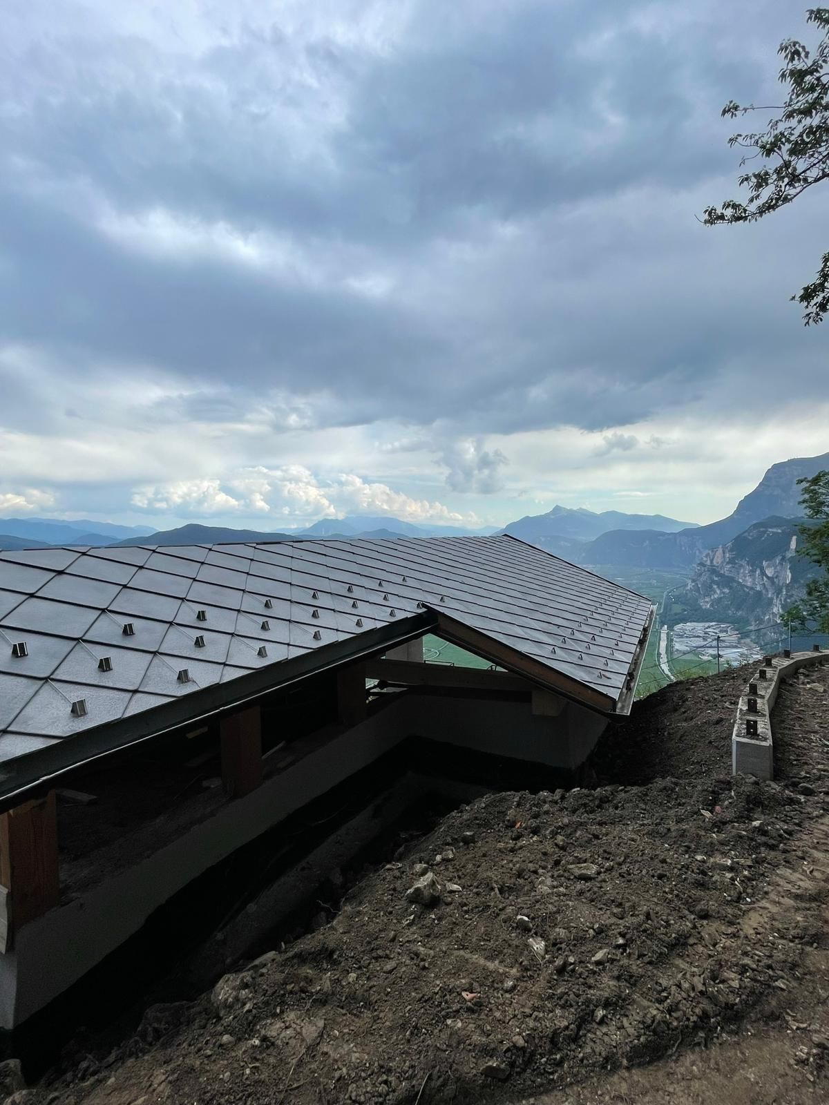 Un edificio in costruzione su un pendio che domina una valle montuosa, con il tetto ricoperto di tegole metalliche.
