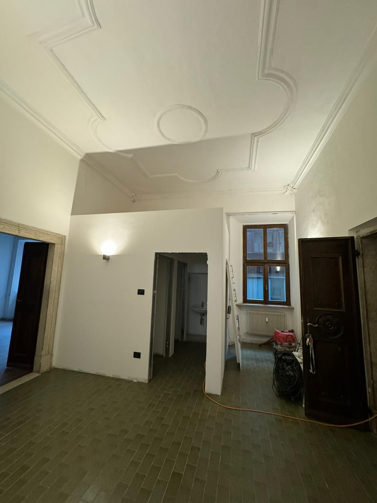 Una stanza con soffitti alti, modanature decorative bianche, pavimento piastrellato, una porta che conduce a un'altra stanza e una porta di legno aperta.