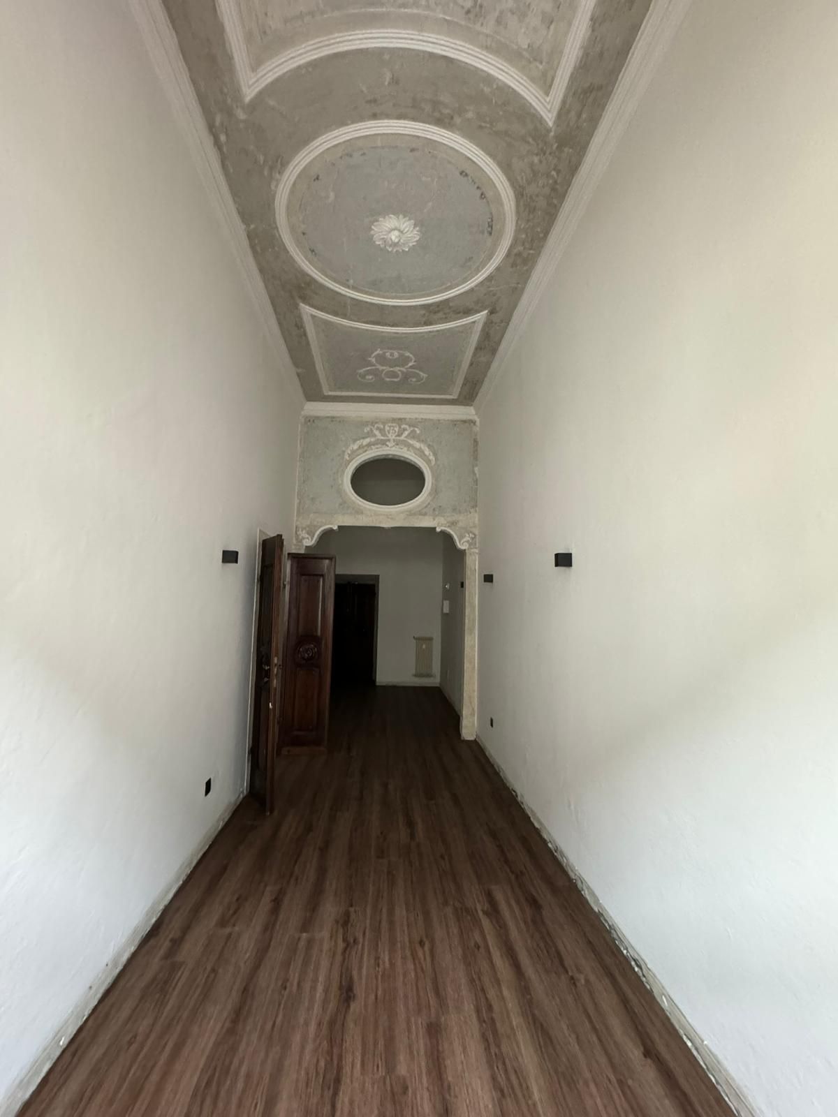 Un corridoio lungo e stretto con pareti bianche, pavimento effetto legno e un soffitto decorato con un elaborato motivo grigio.