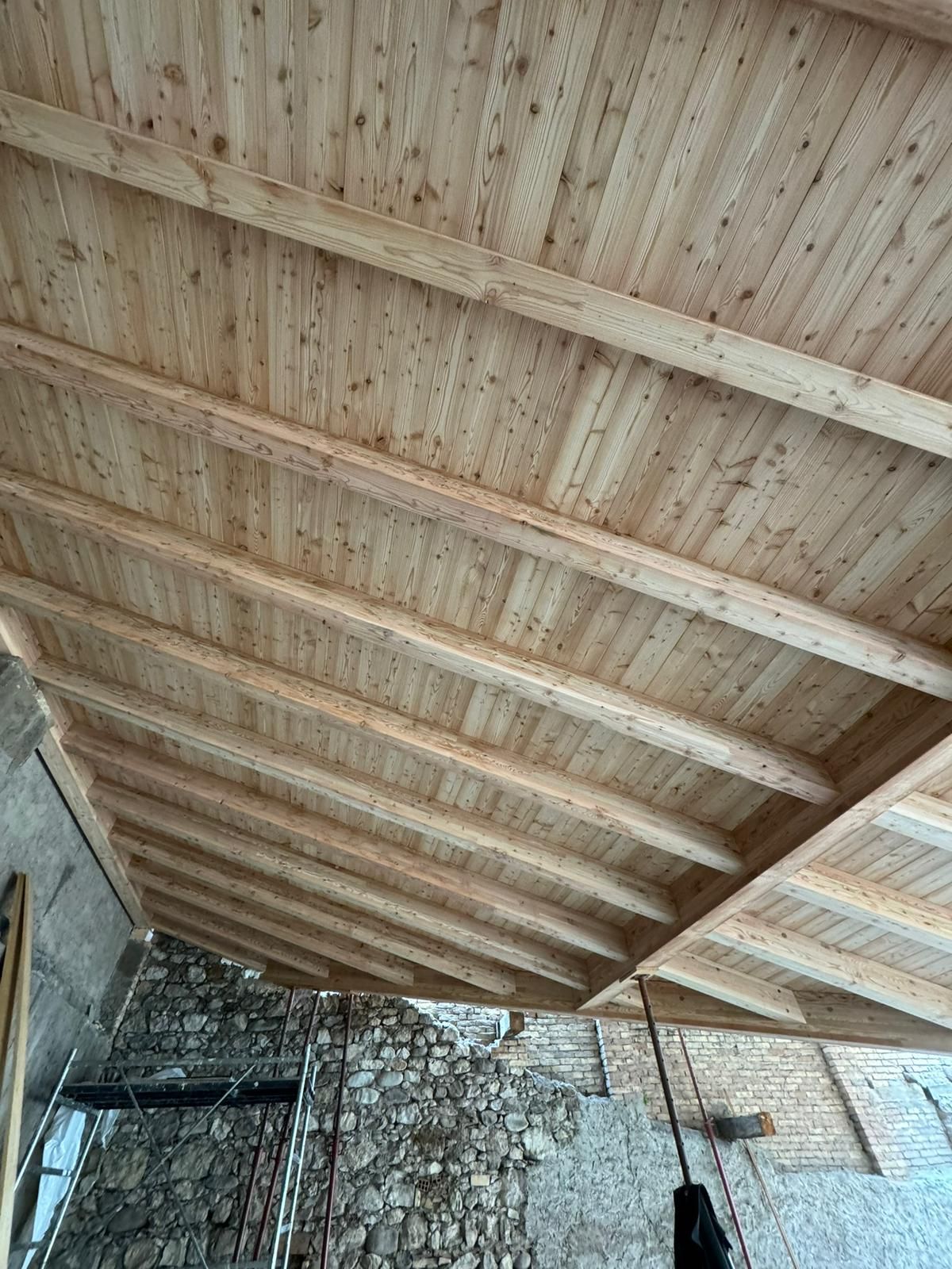 Vista dal basso di una struttura di sostegno in legno di nuova costruzione, composta da soffitto e tetto, sopra un muro di pietra.