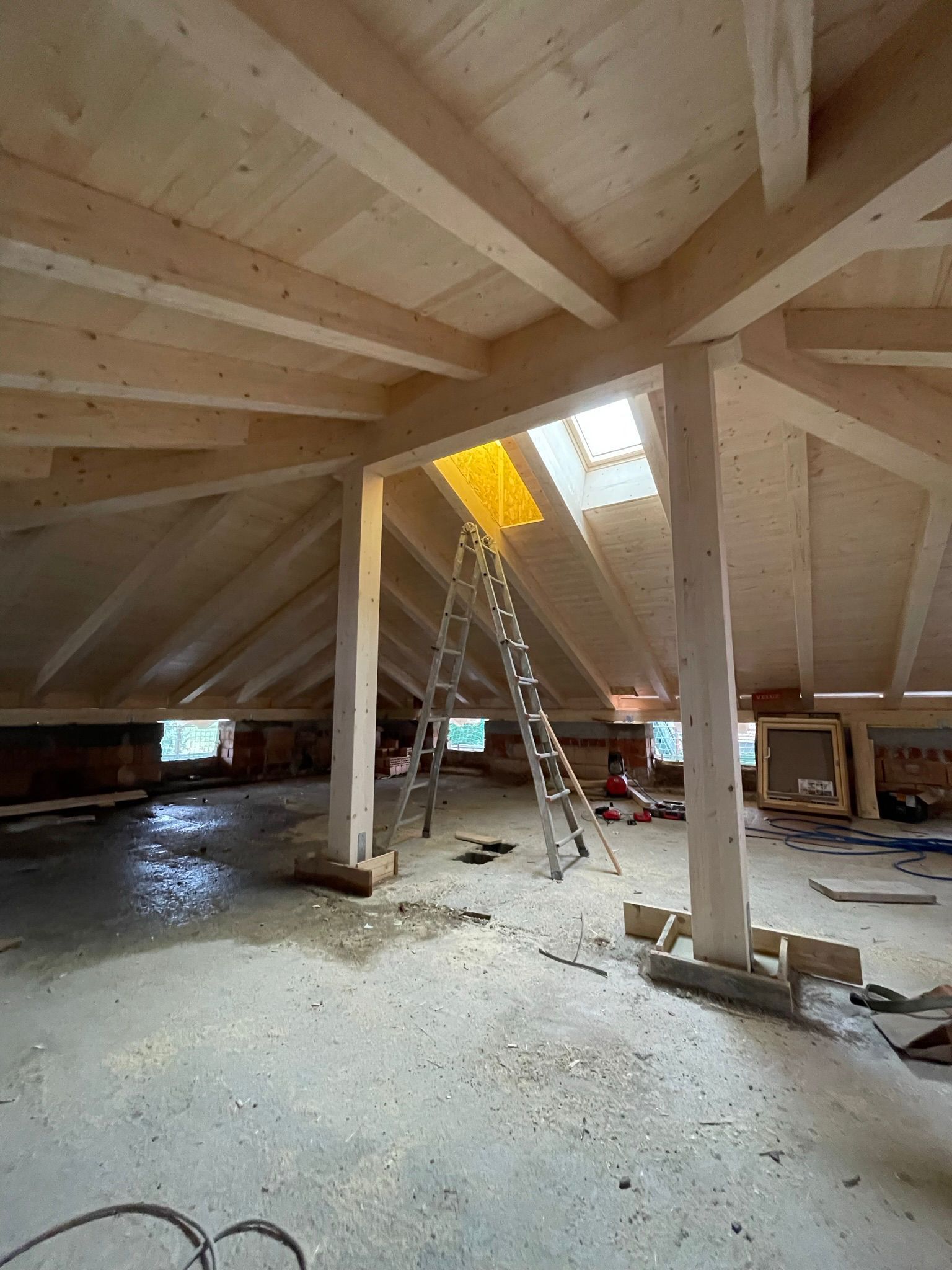 In una soffitta non rifinita, con travi di legno a vista e pavimento in cemento, una scala è posizionata sotto un lucernario.