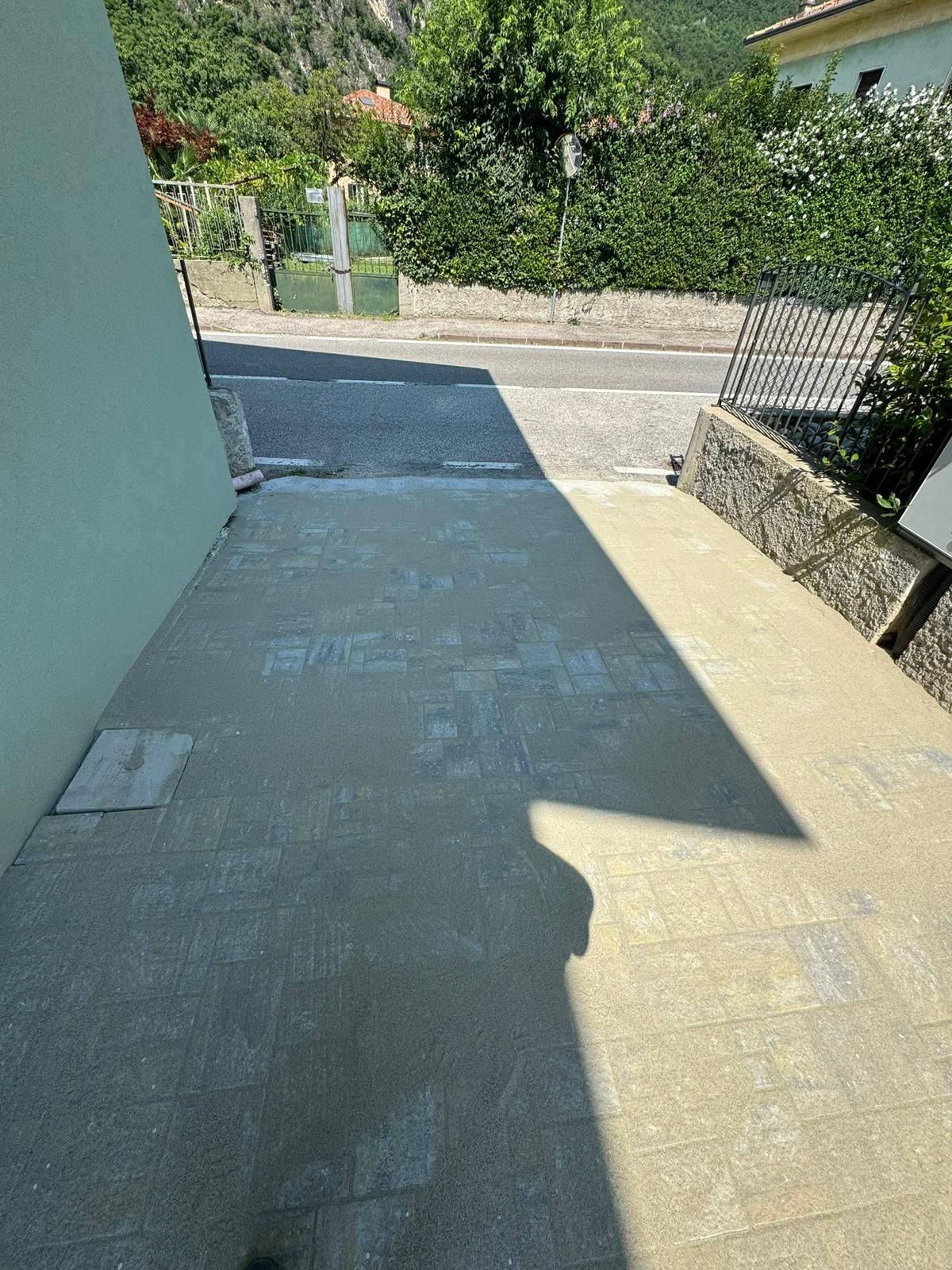 Un sentiero lastricato con pietre grigie e beige chiaro che conduce a una strada, parzialmente nascosta da un'ombra scura di forma triangolare.