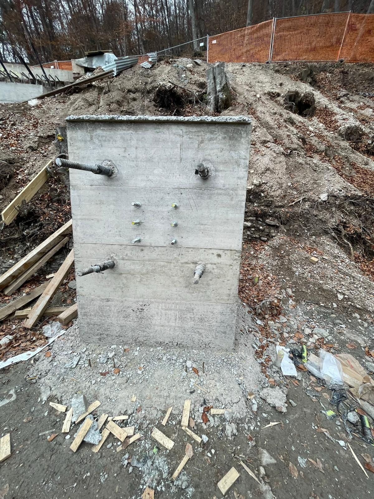 Un blocco di cemento in un cantiere edile, con bulloni metallici sporgenti, incastrato nel terreno vicino a una recinzione di sicurezza arancione.