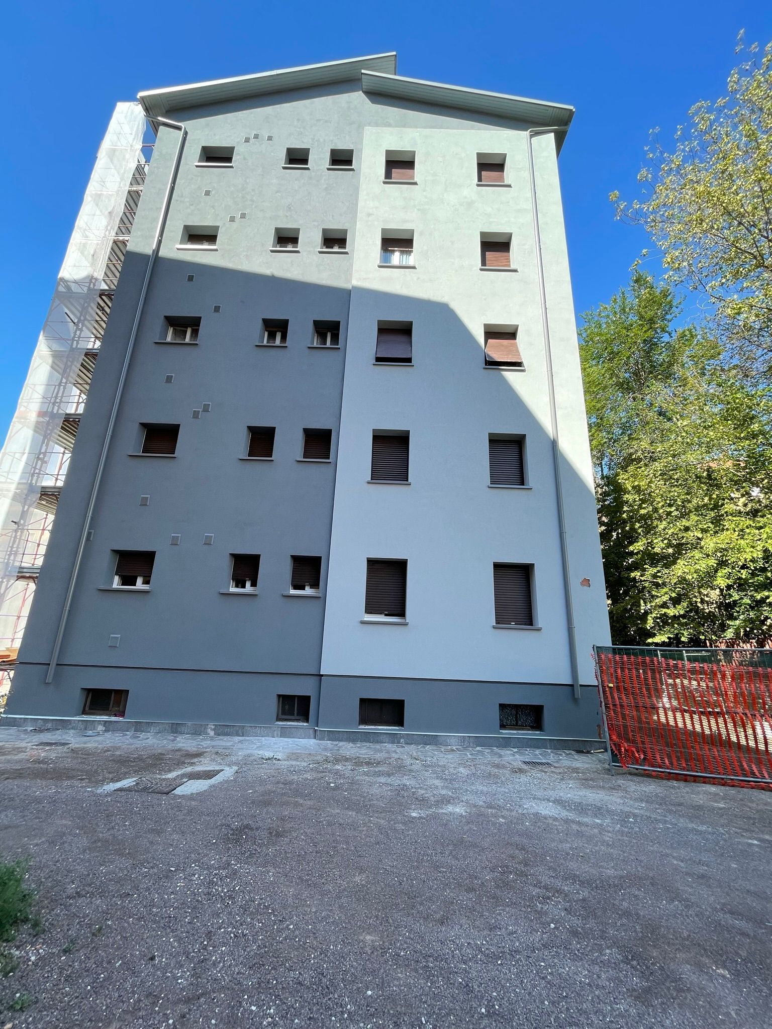 Un condominio a più piani con facciata grigia e grigio chiaro e un cortile anteriore con ciottoli, sotto un cielo azzurro e limpido.
