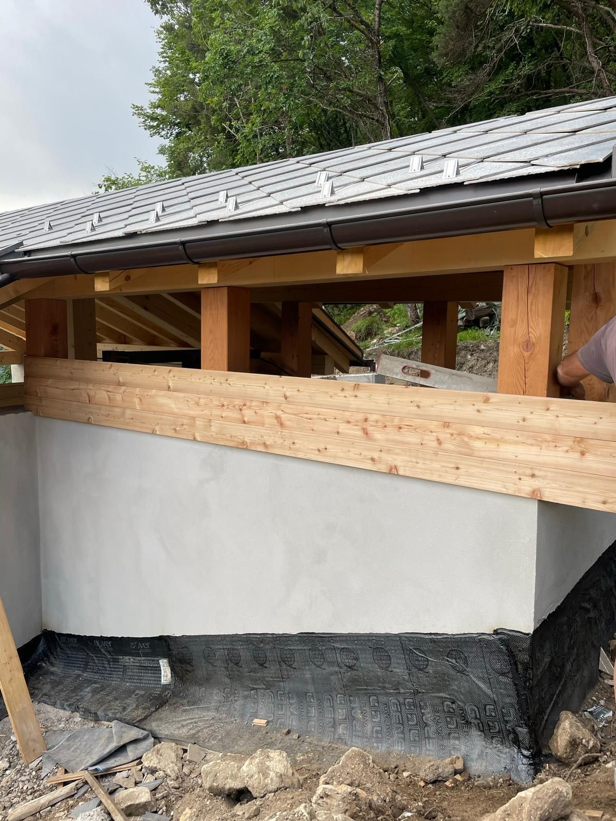 Una struttura in legno parzialmente costruita, con una base in cemento grigio chiaro, si erge sotto un tetto di tegole di ardesia grigia.