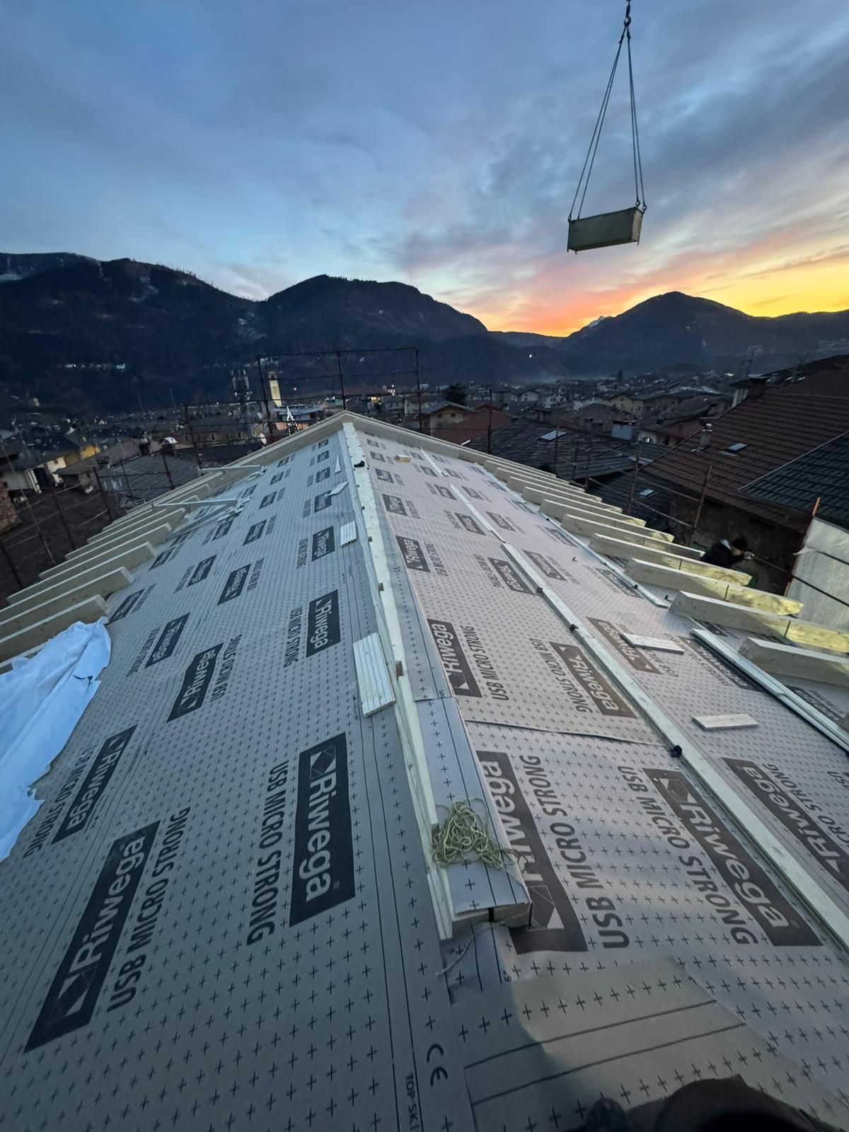 Una veduta grandangolare di un tetto in costruzione al tramonto, con un carico sollevato da una gru sospeso contro il profilo montuoso.