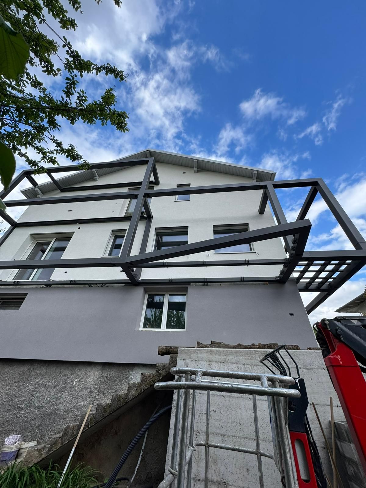 Una casa a due piani sotto un cielo azzurro e nuvoloso, caratterizzata da una struttura metallica grigia non rifinita annessa alla facciata esterna.