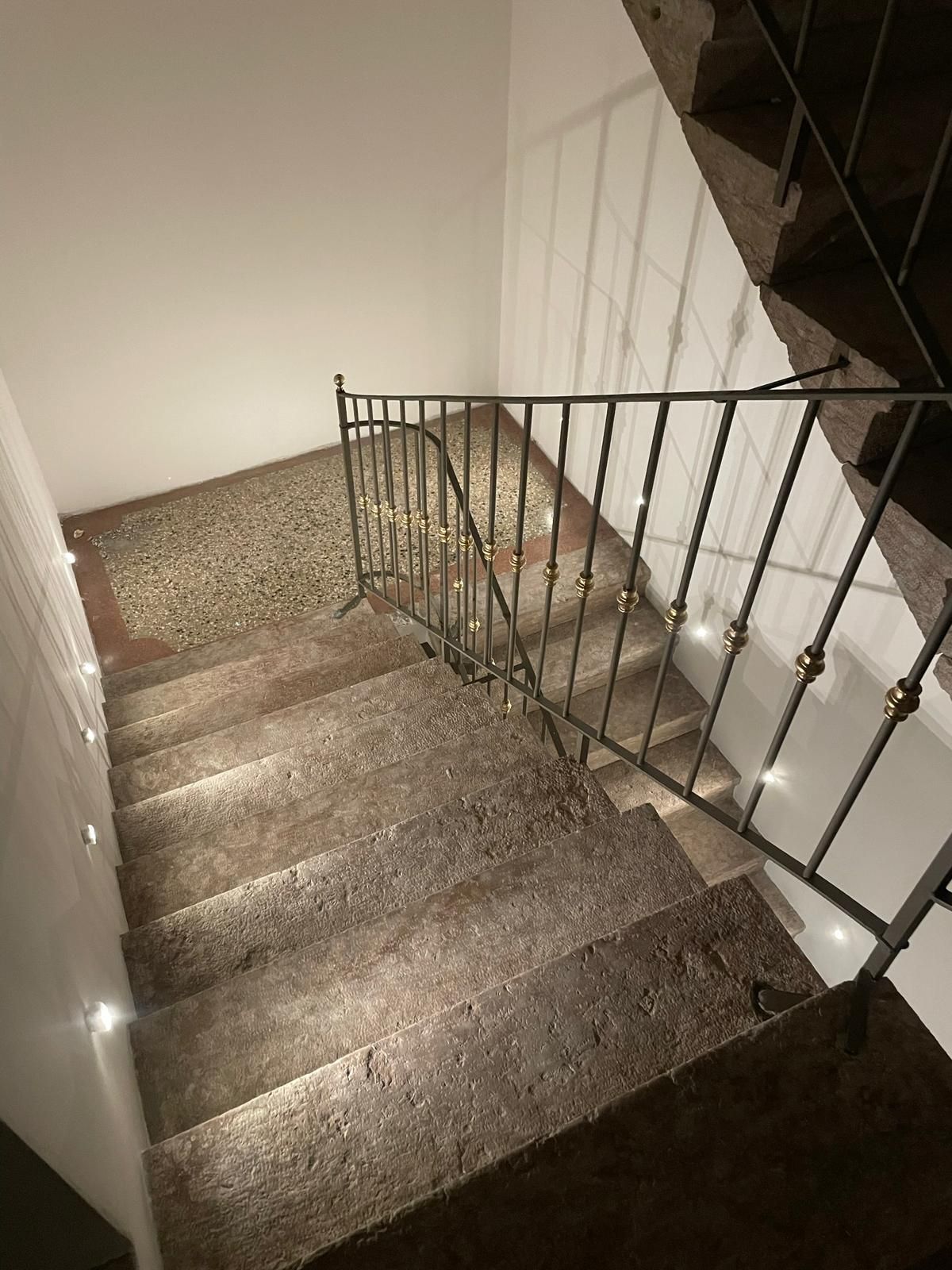 Una scala in pietra con ringhiera in ferro, illuminata da piccole applique a parete, che conduce a un pianerottolo.