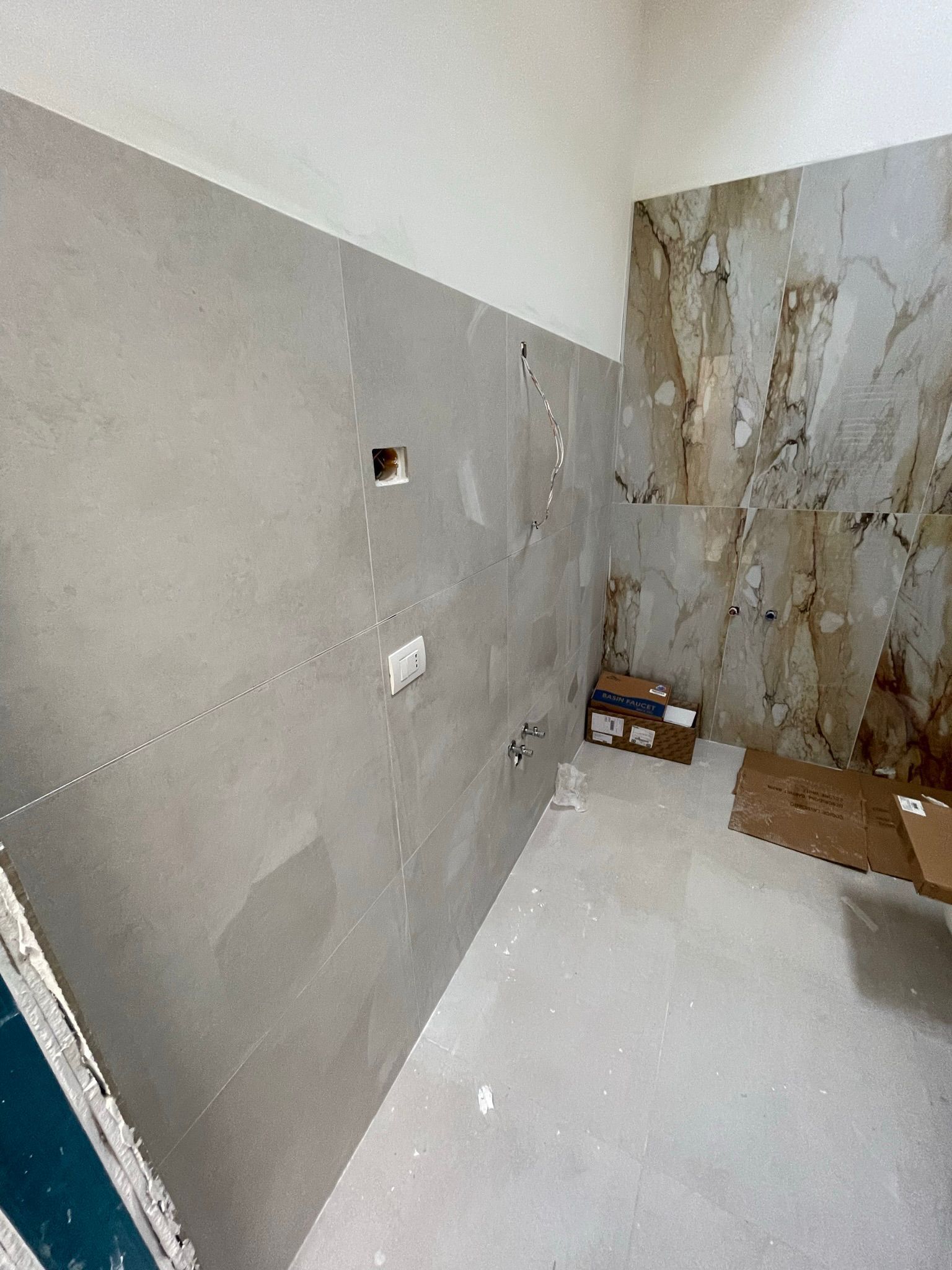 Una stanza in fase di ristrutturazione con pareti rivestite di piastrelle grigie a sinistra, una parete con rivestimento effetto pietra a destra e pavimento grezzo.