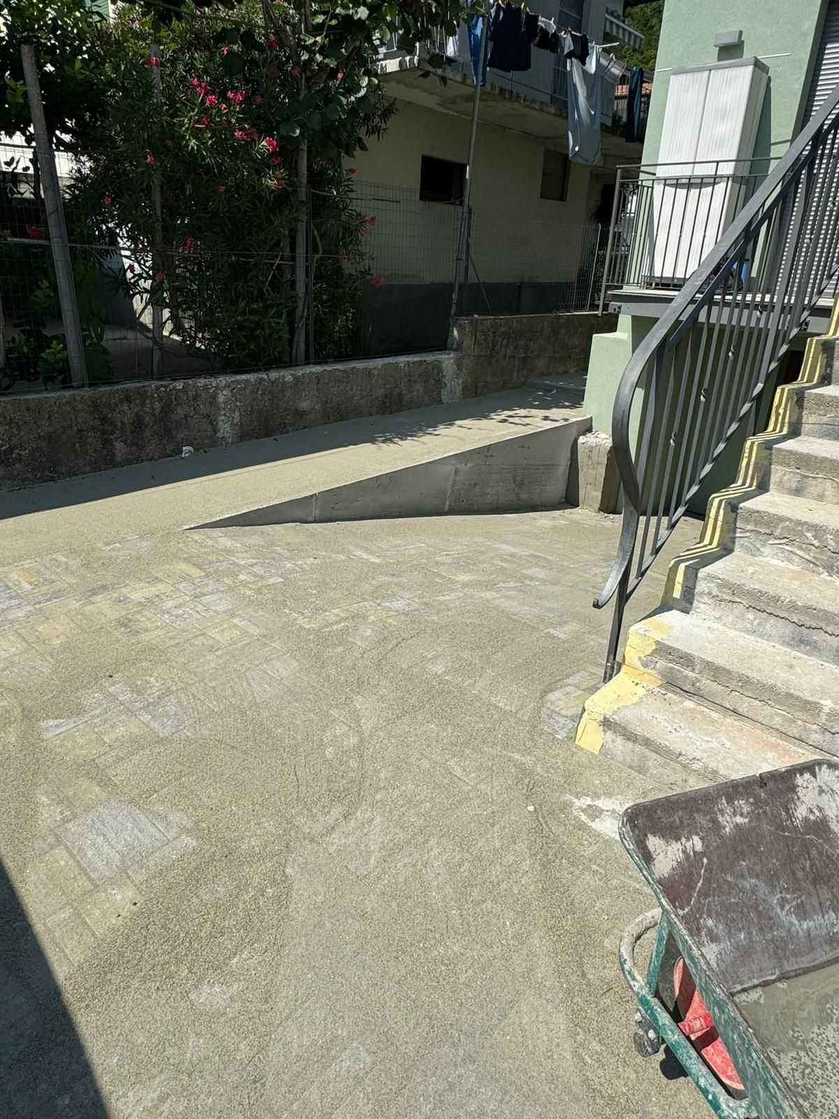 Una rampa in cemento che conduce all'ingresso di un'abitazione, situata accanto a una scala con ringhiera metallica.