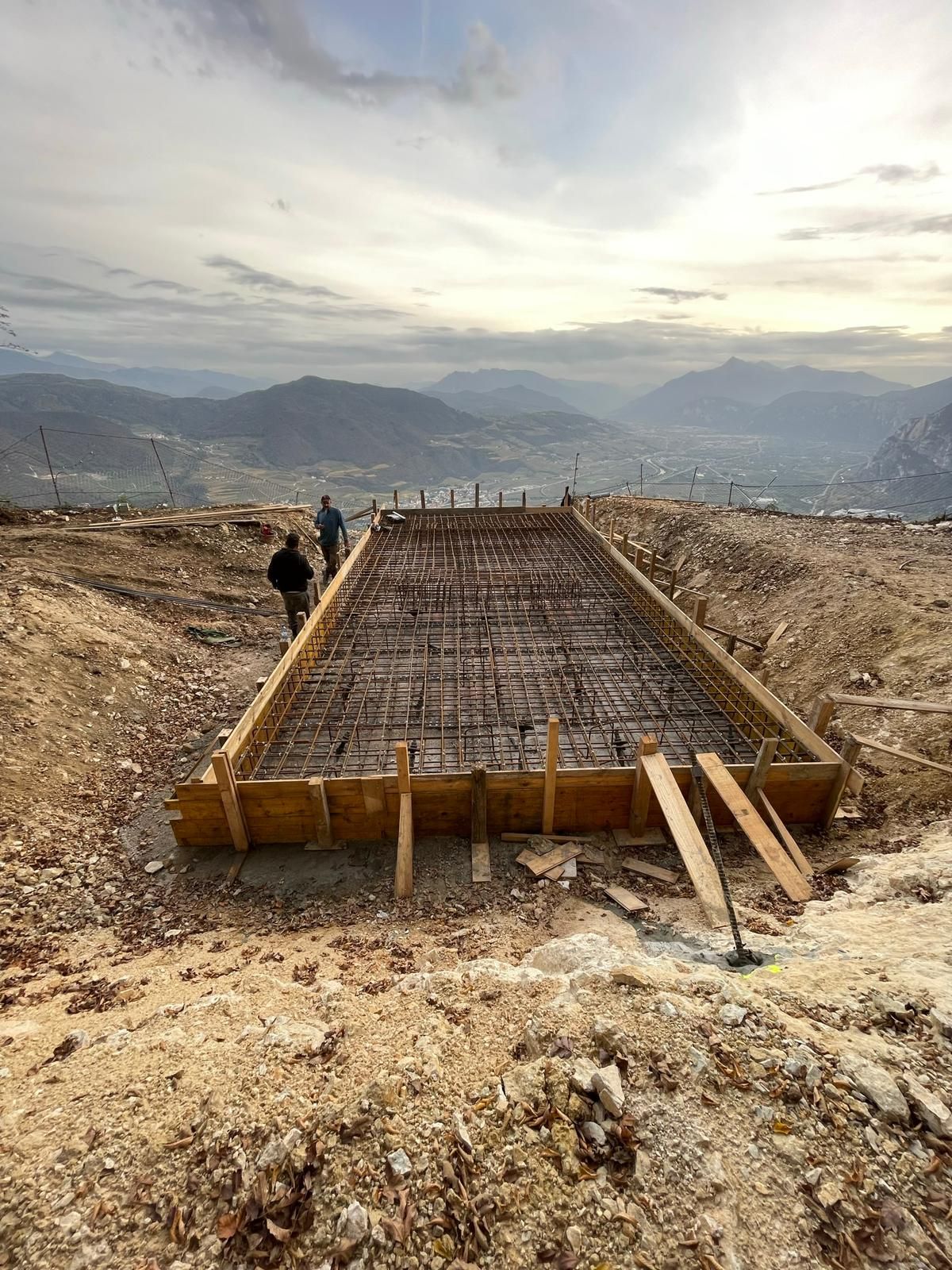 Due persone sono in piedi accanto a una cassaforma rettangolare in cemento armato, situata su un fianco di montagna sotto un cielo nuvoloso.