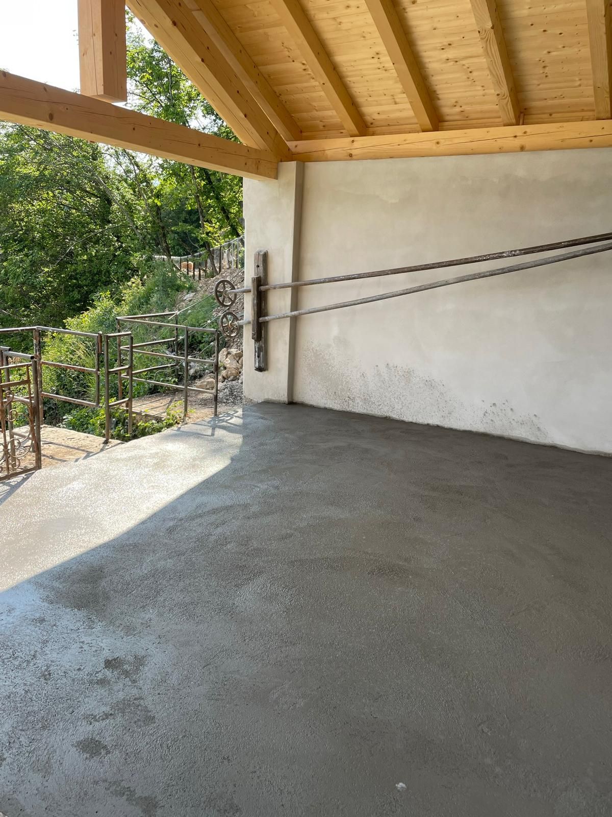 Un patio esterno coperto con pavimento in cemento grigio appena gettato, tetto con travi in ​​legno e parete beige con cavi d'acciaio.