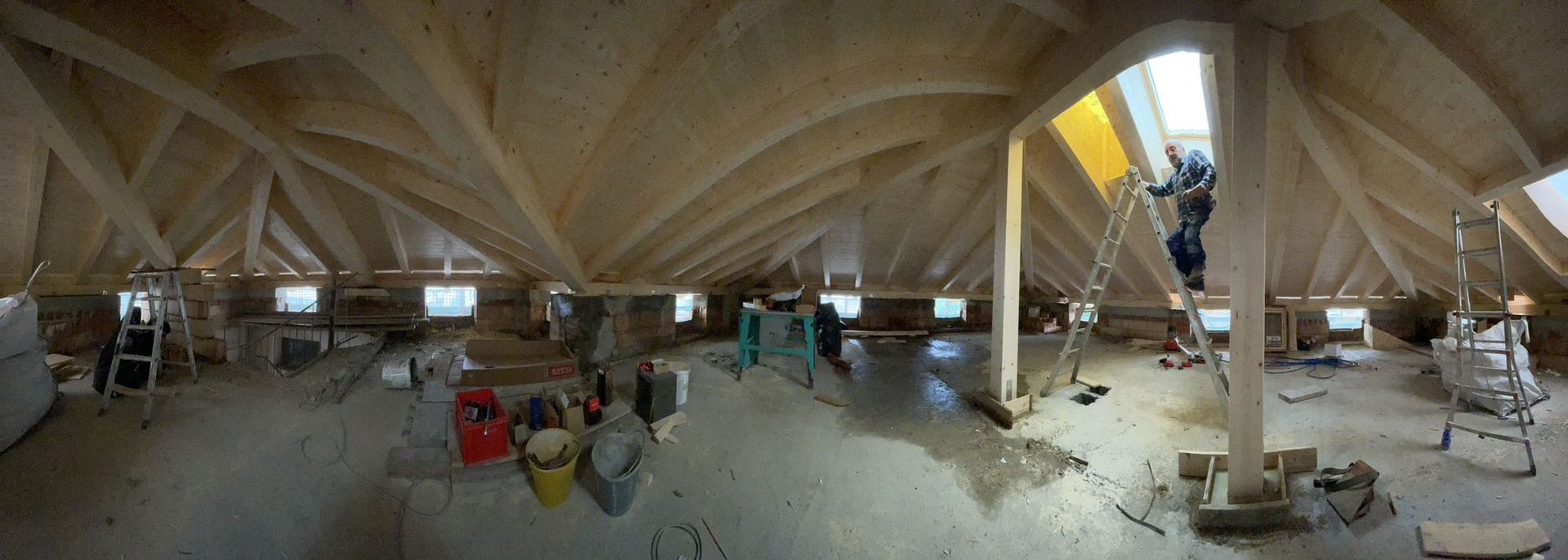 Veduta grandangolare degli interni di una soffitta in costruzione, con travi di legno a vista e un operaio su una scala.