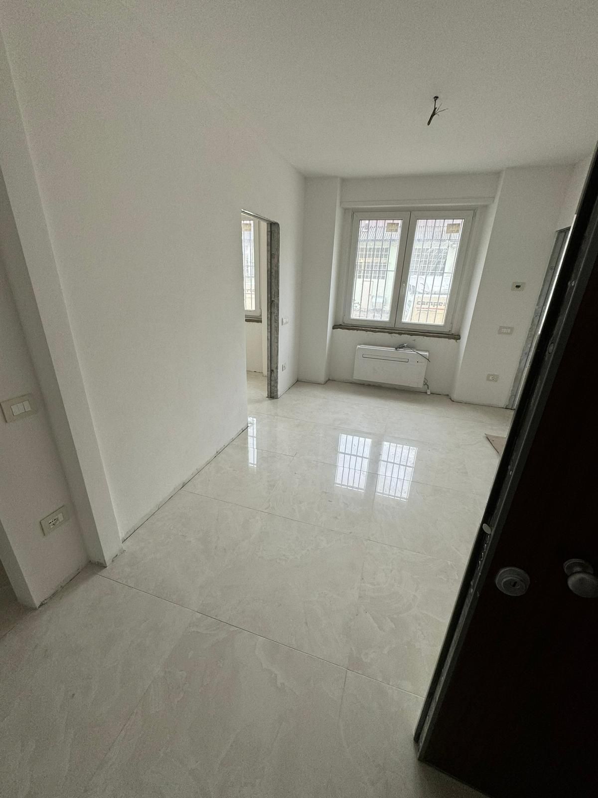 Una stanza luminosa e vuota con pareti bianche, pavimento in piastrelle chiare e lucide e una finestra che lascia entrare la luce naturale.