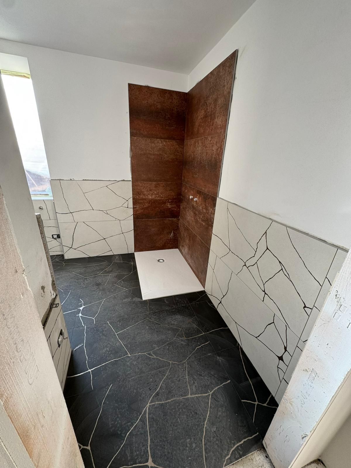 Un bagno in fase di ristrutturazione con pavimento in piastrelle a effetto craquelé bianche e nere, piastrelle bianche alle pareti e piastrelle marroni nella doccia.