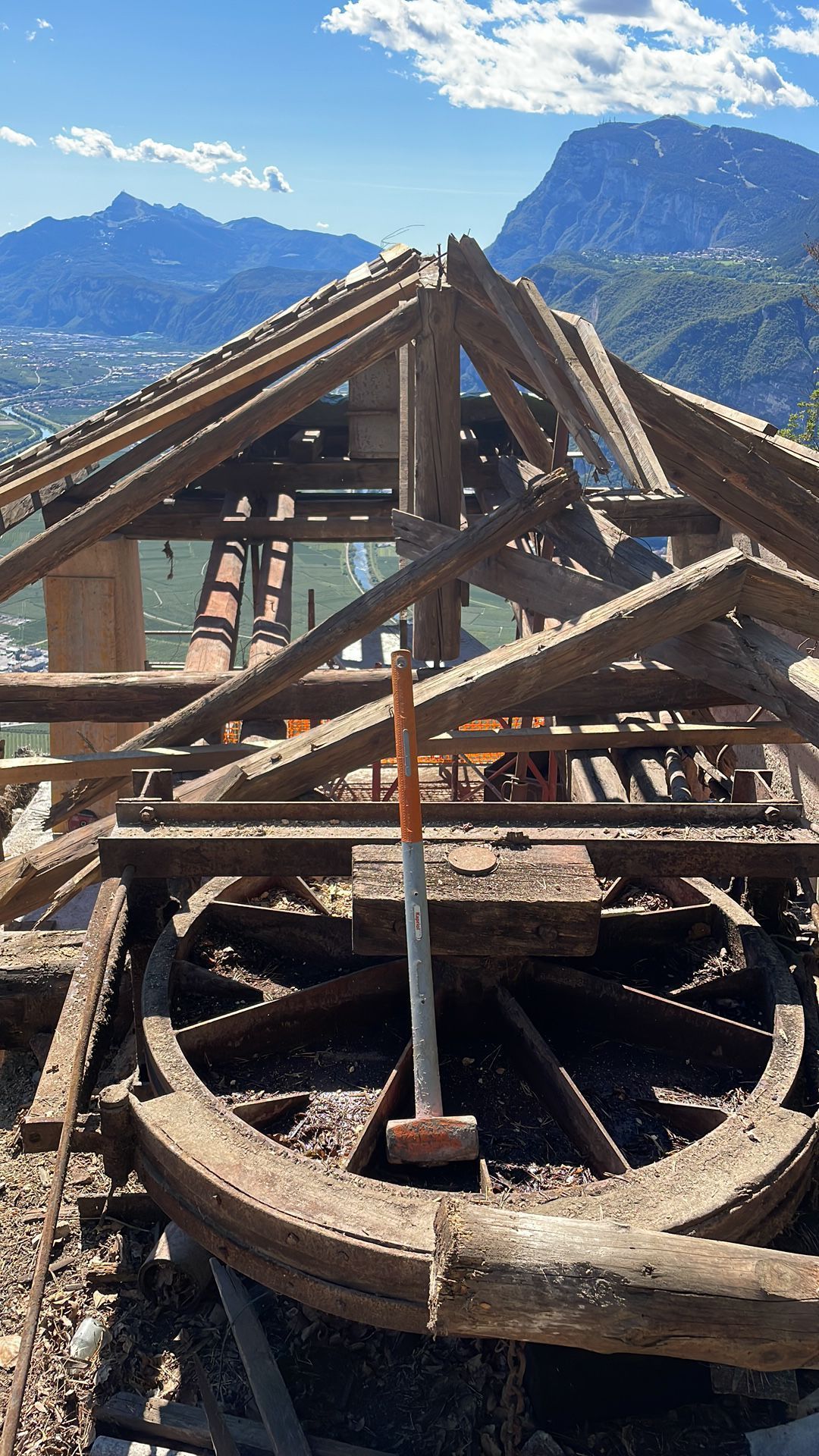 Una mazza poggia su una grande ruota di legno all'interno di un'antica struttura in legno, sullo sfondo di un paesaggio montuoso e soleggiato.