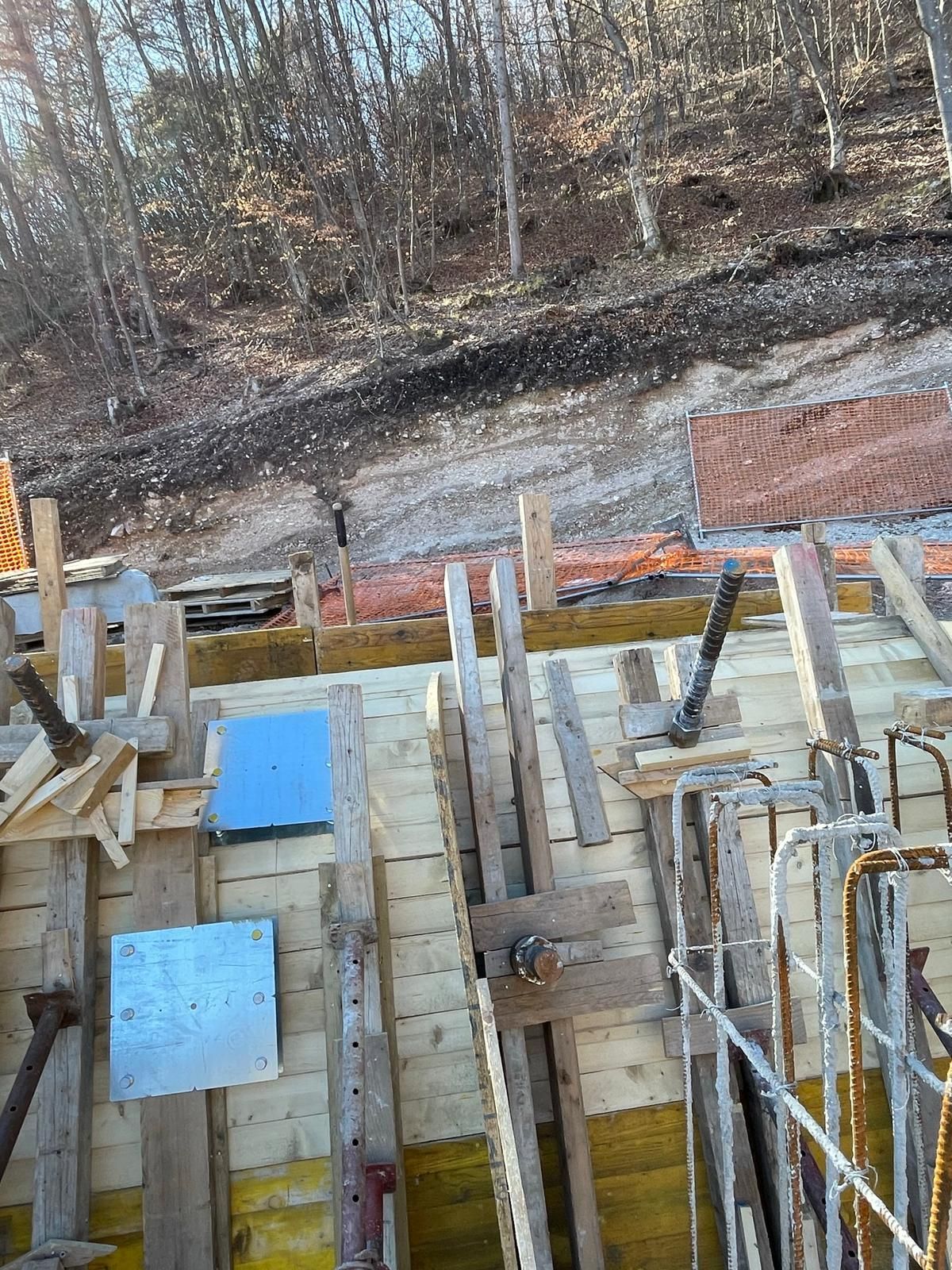 Cantiere edile con casseforme in cemento, travi di supporto metalliche e armature in barre d'armatura, addossato a un pendio boscoso.