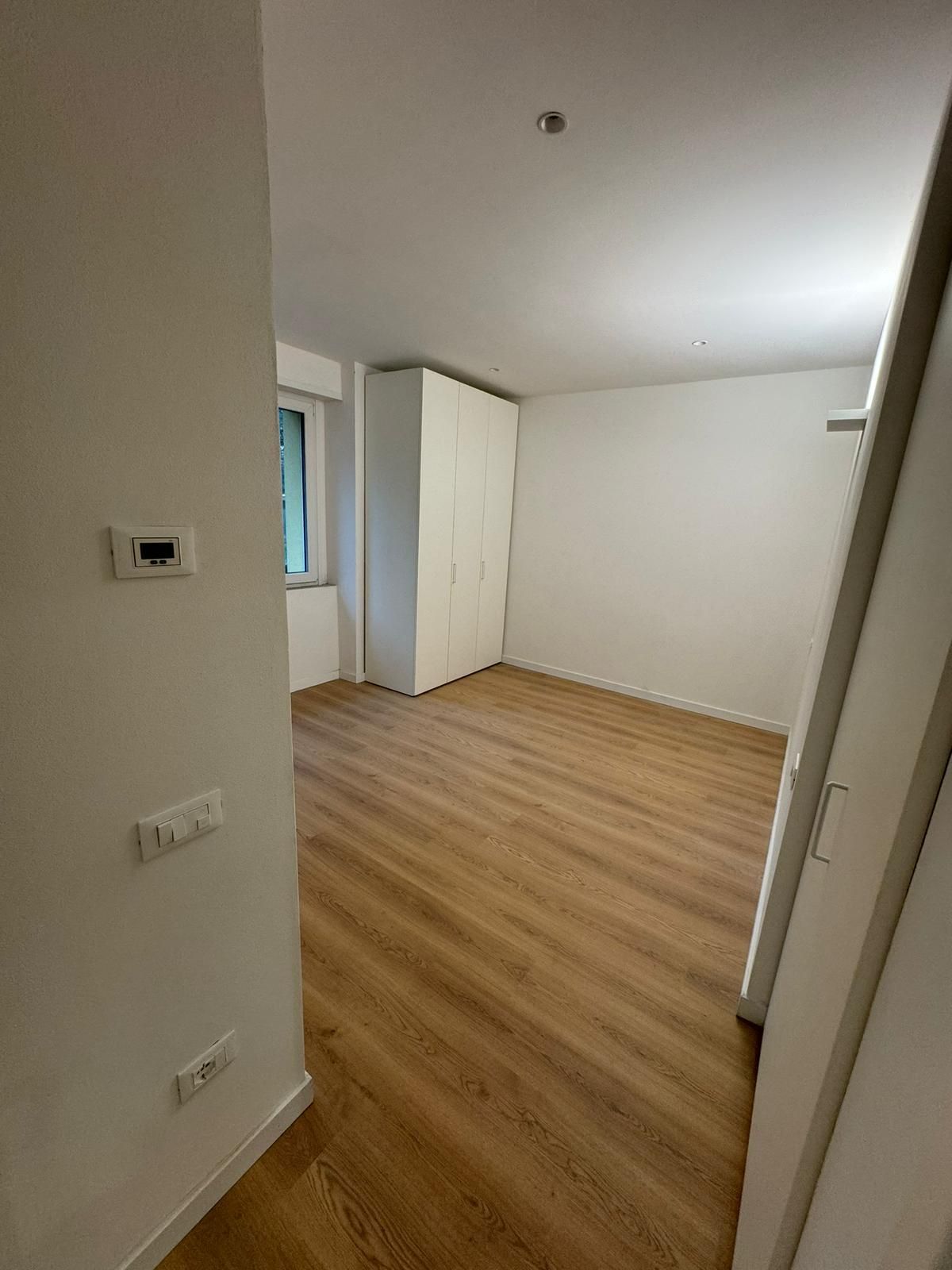 Una stanza luminosa e vuota con pavimento in legno chiaro, pareti bianche e un alto armadio bianco in un angolo.