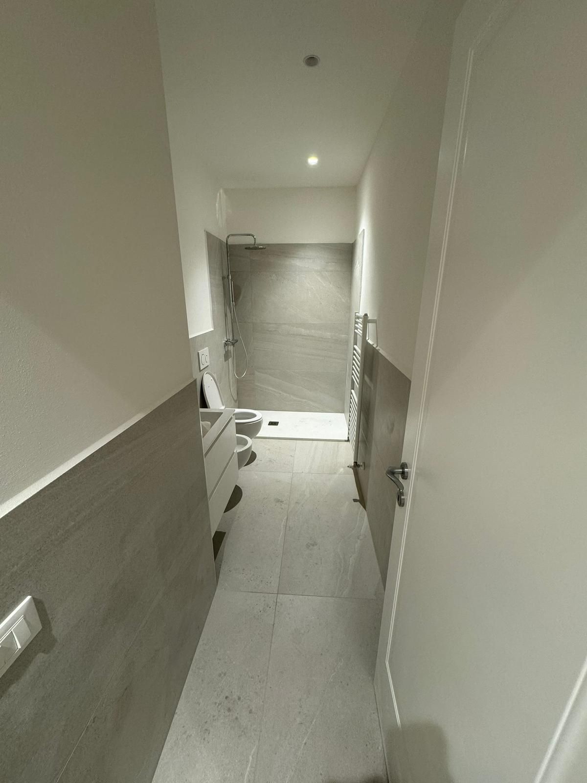 Un bagno stretto e moderno con piastrelle grigie a parete e pavimento, un mobile lavabo bianco, un bidet e una cabina doccia in fondo.